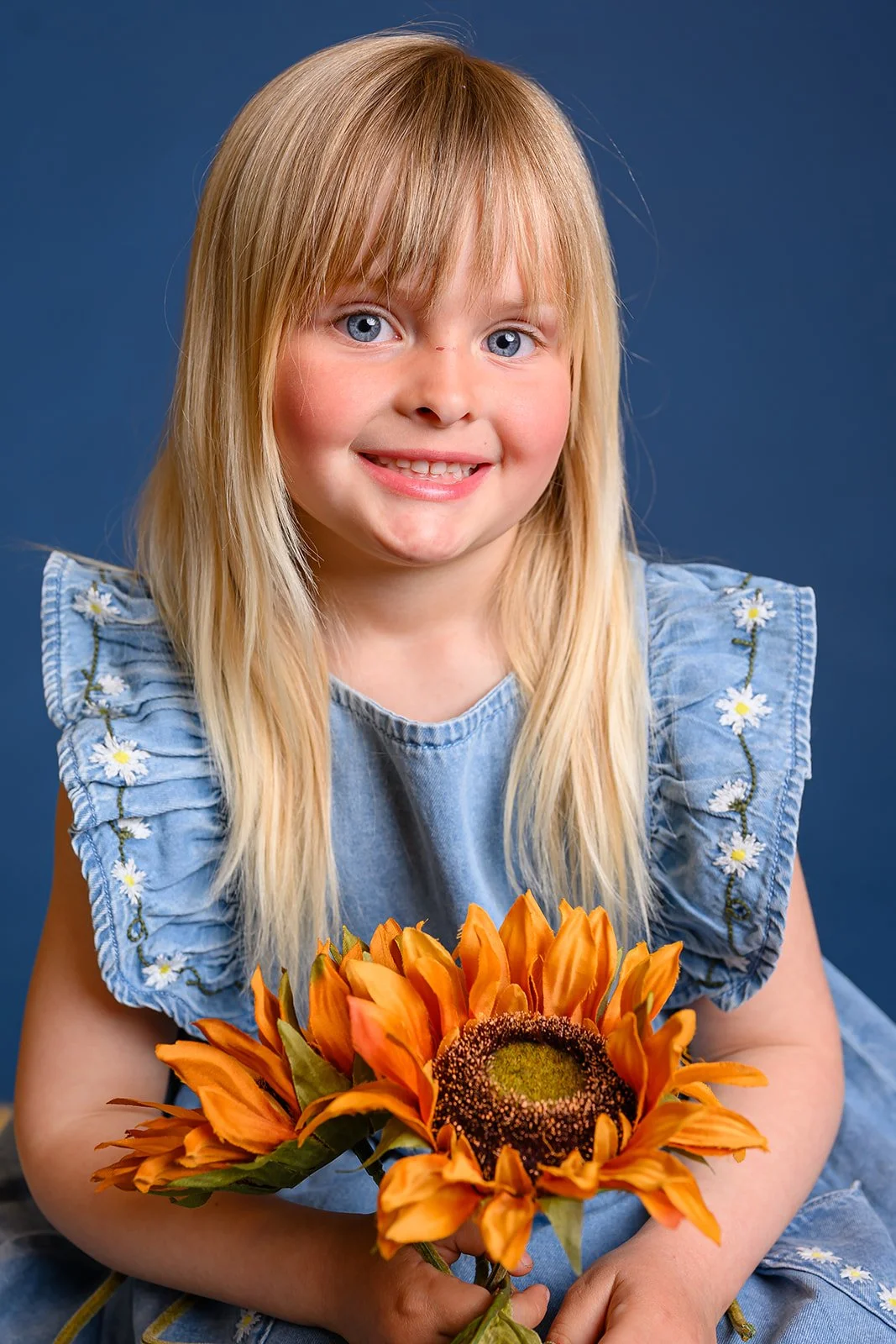Smilende blond jente i blomstrete blå kjole holder en bukett med oransje solsikker foran blå bakgrunn.