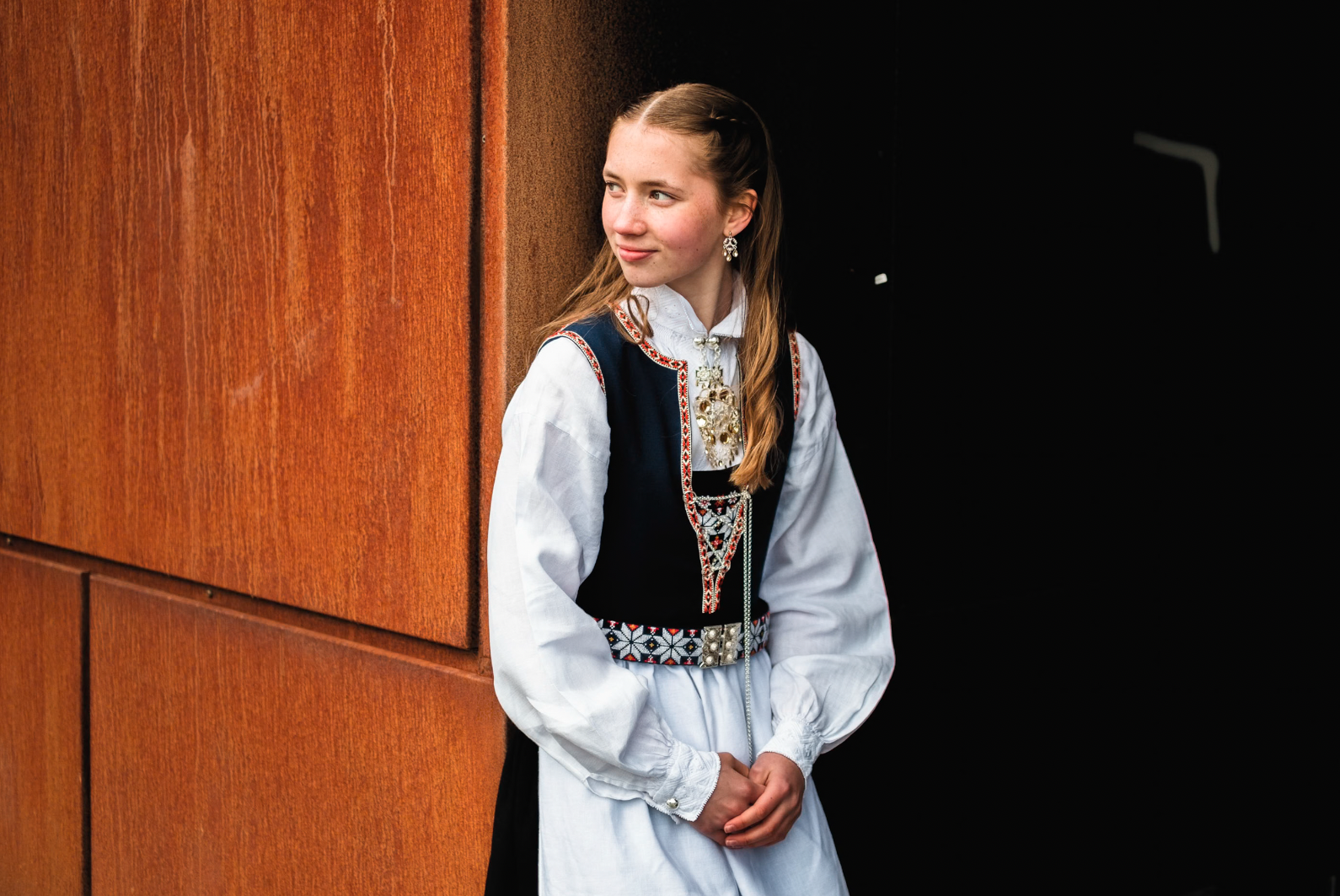 Ung kvinne i tradisjonell norsk bunad står mot en vegg av rustent metall.