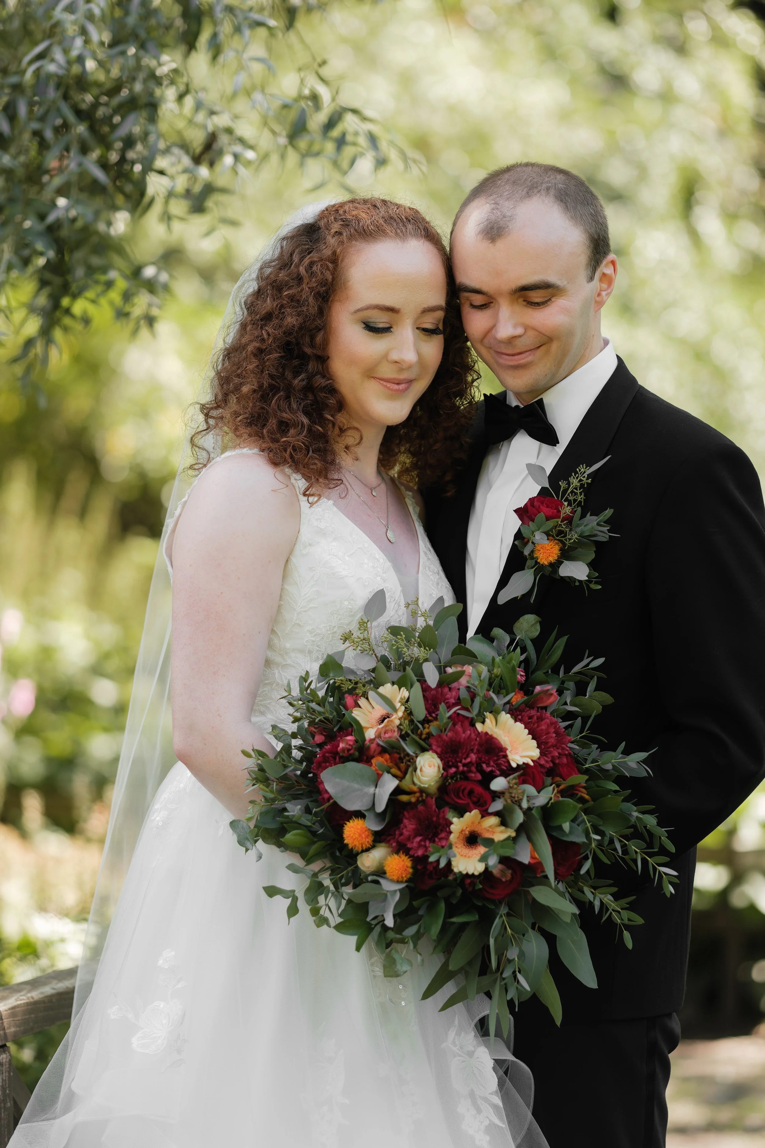 Et nygift brudepar står sammen, kvinnen med en hvit brudekjole og mannen i svart dress med sløyfe, de holder en stor blomsterbukett, omgitt av grønt blader, i en utendørs setting.