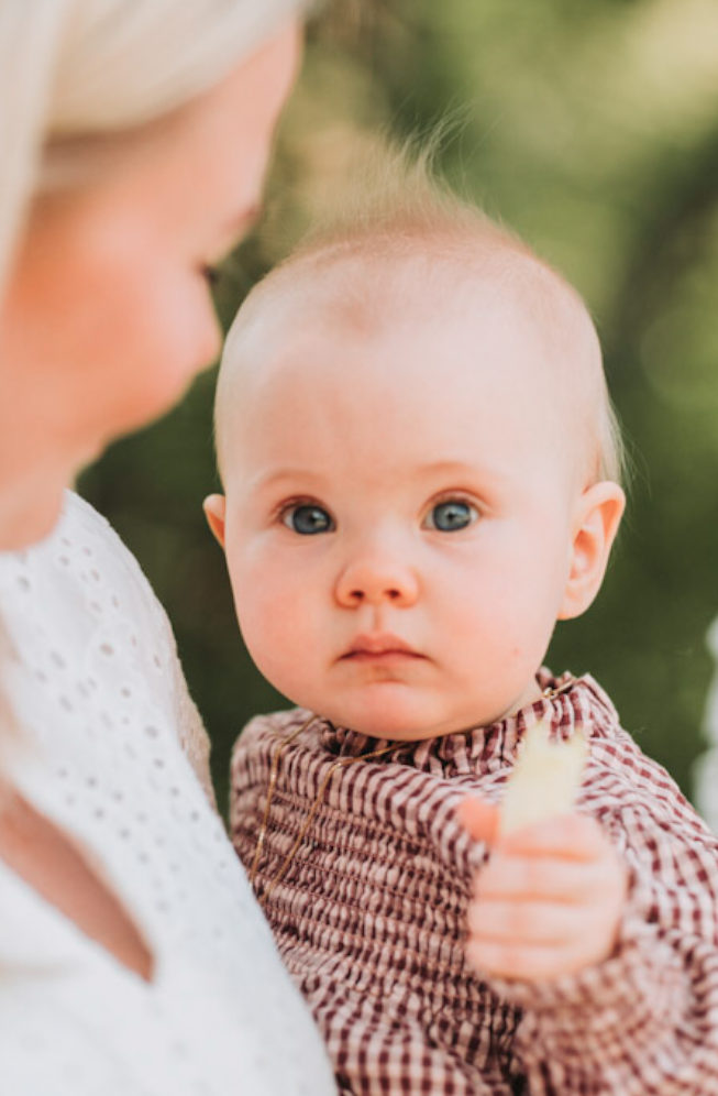 En baby blir holdt av en person, sannsynligvis en mor, i utendørsmiljø med grønt bakgrunn, babyen har blå øyne og lyse hår, og holder en popsicle.