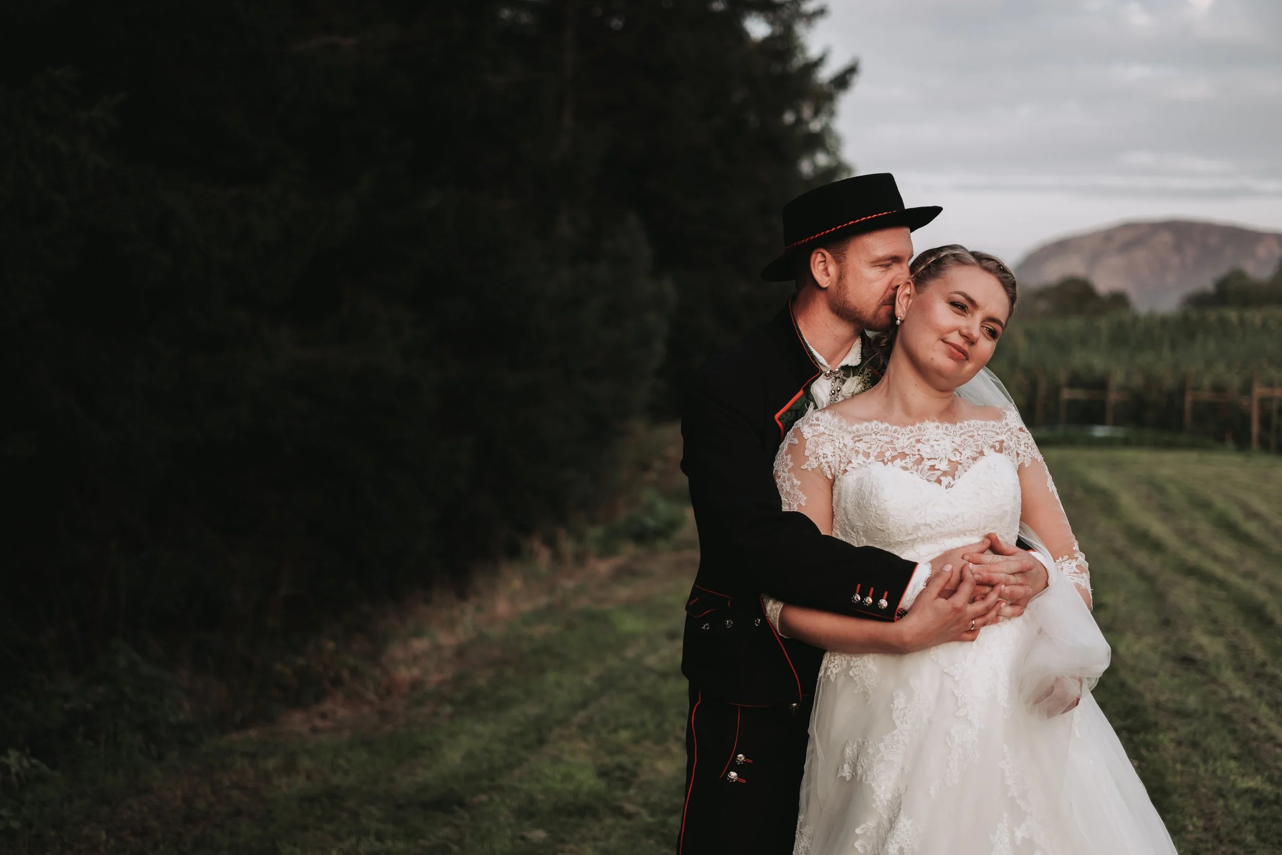 Et brudepar står i en grønn utendørssetting, mannen i tradisjonell svart og rød dress med hatt, kvinnen i hvit brudekjole, de holder hender og nyter øyeblikket.