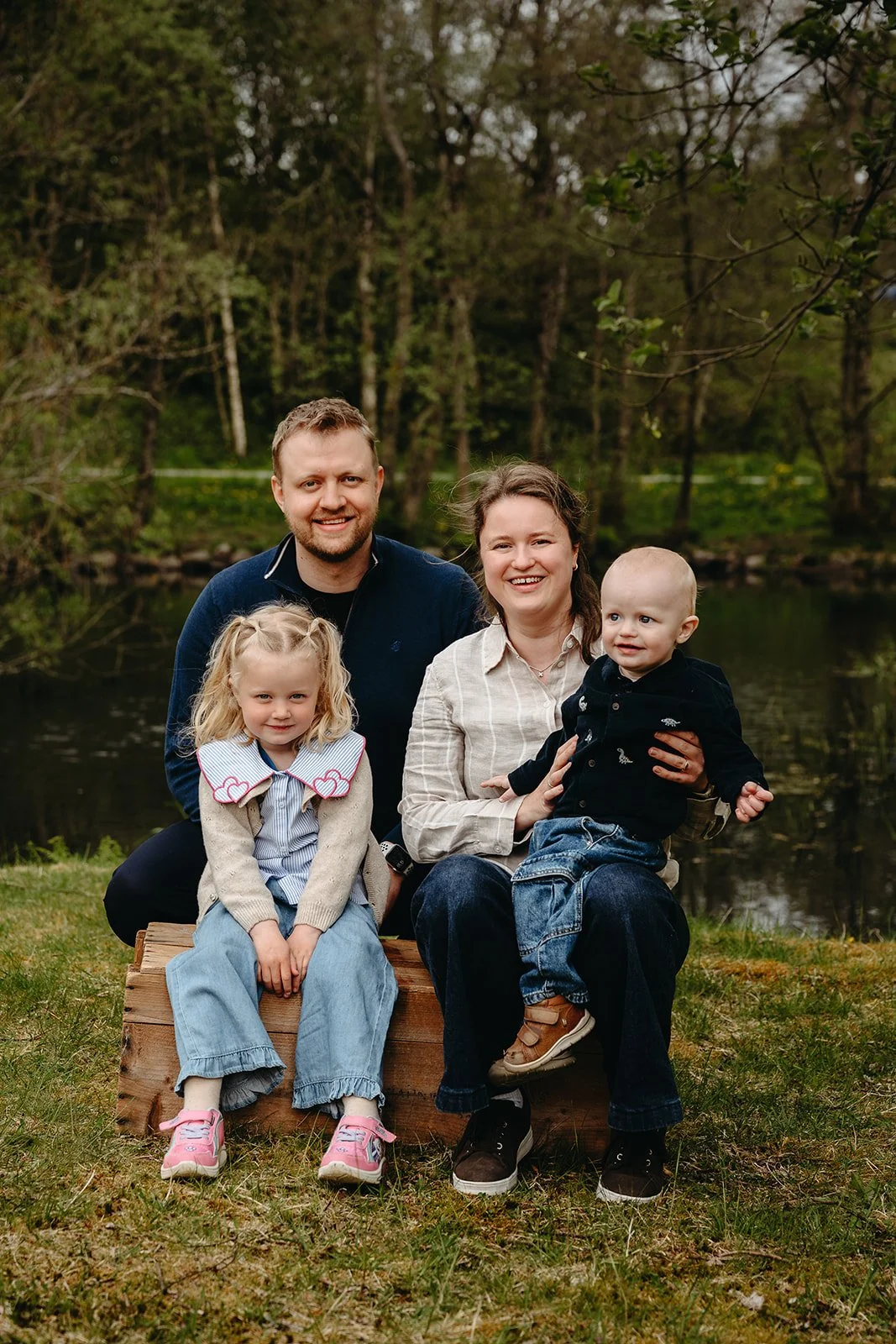 Familie på fire sitter på en trebenk i en skog ved en liten innsjø.