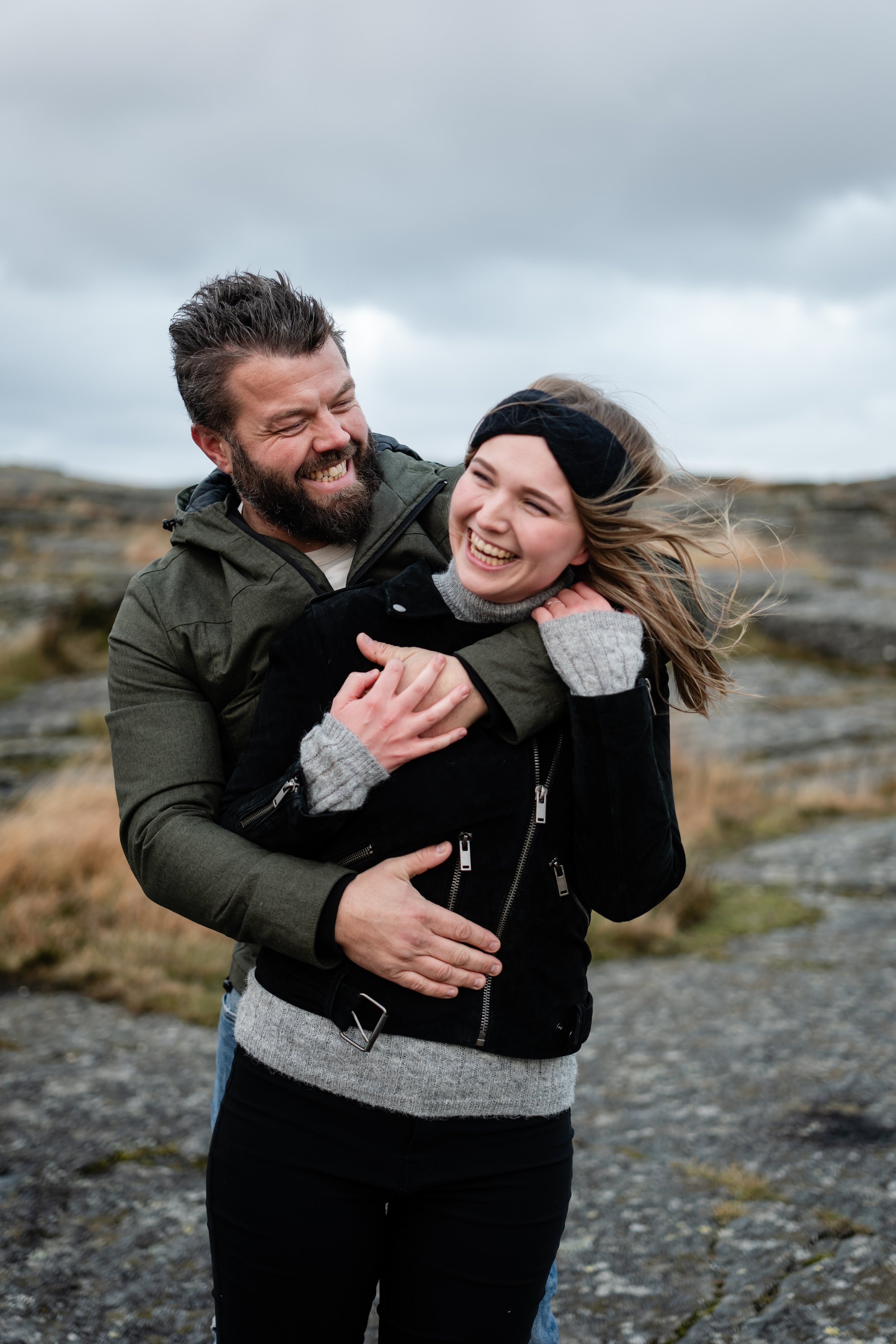 Et par som ler og klemmer hverandre utendørs i vindfullt vær, med overskyet himmel og landskap av steiner og grashøyder i bakgrunnen.