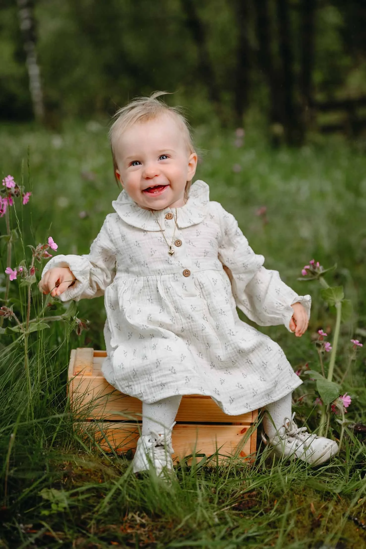 Smilende baby i hvit kjole sitter på en trekasse omgitt av markblomster i skogen.