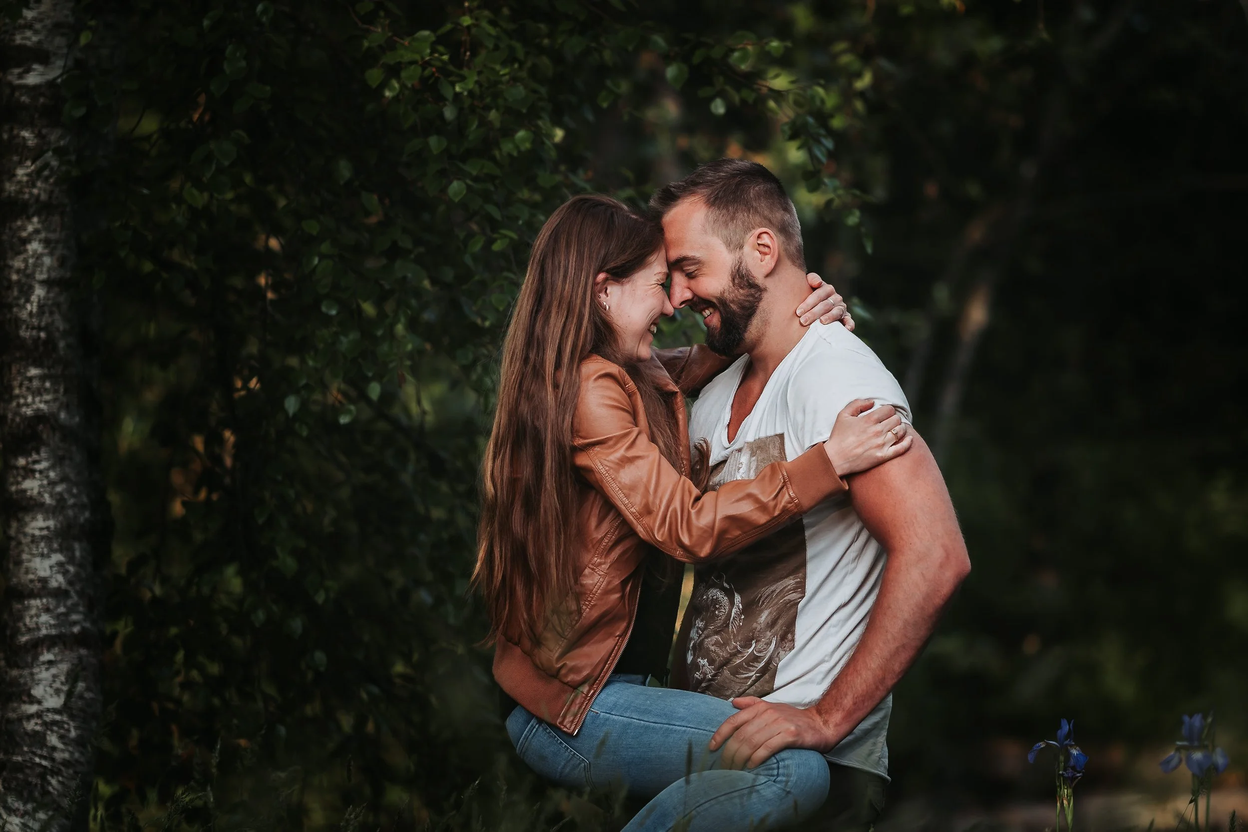 Et ungt par, begge med smilende ansikter, går tett sammen i en park om kvelden, omgitt av grønne blader og blå blomster.