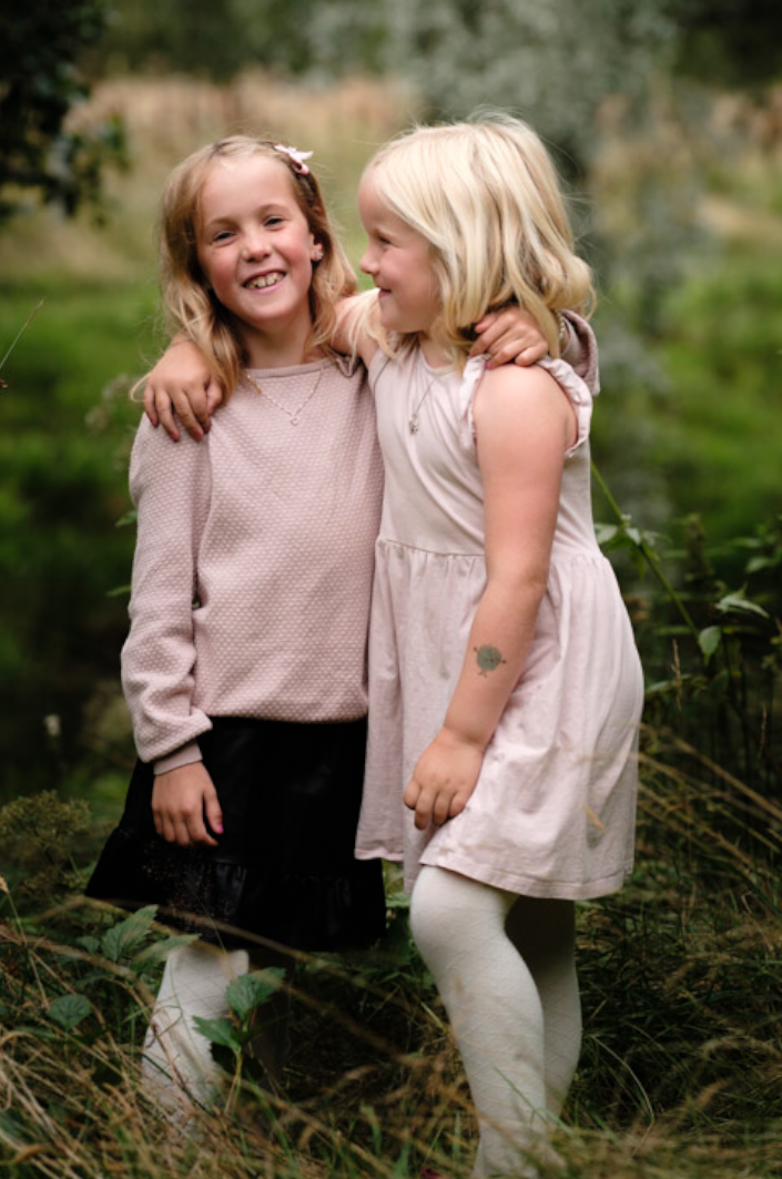 To unge jenter i lyse klær står arm i arm i en skog.