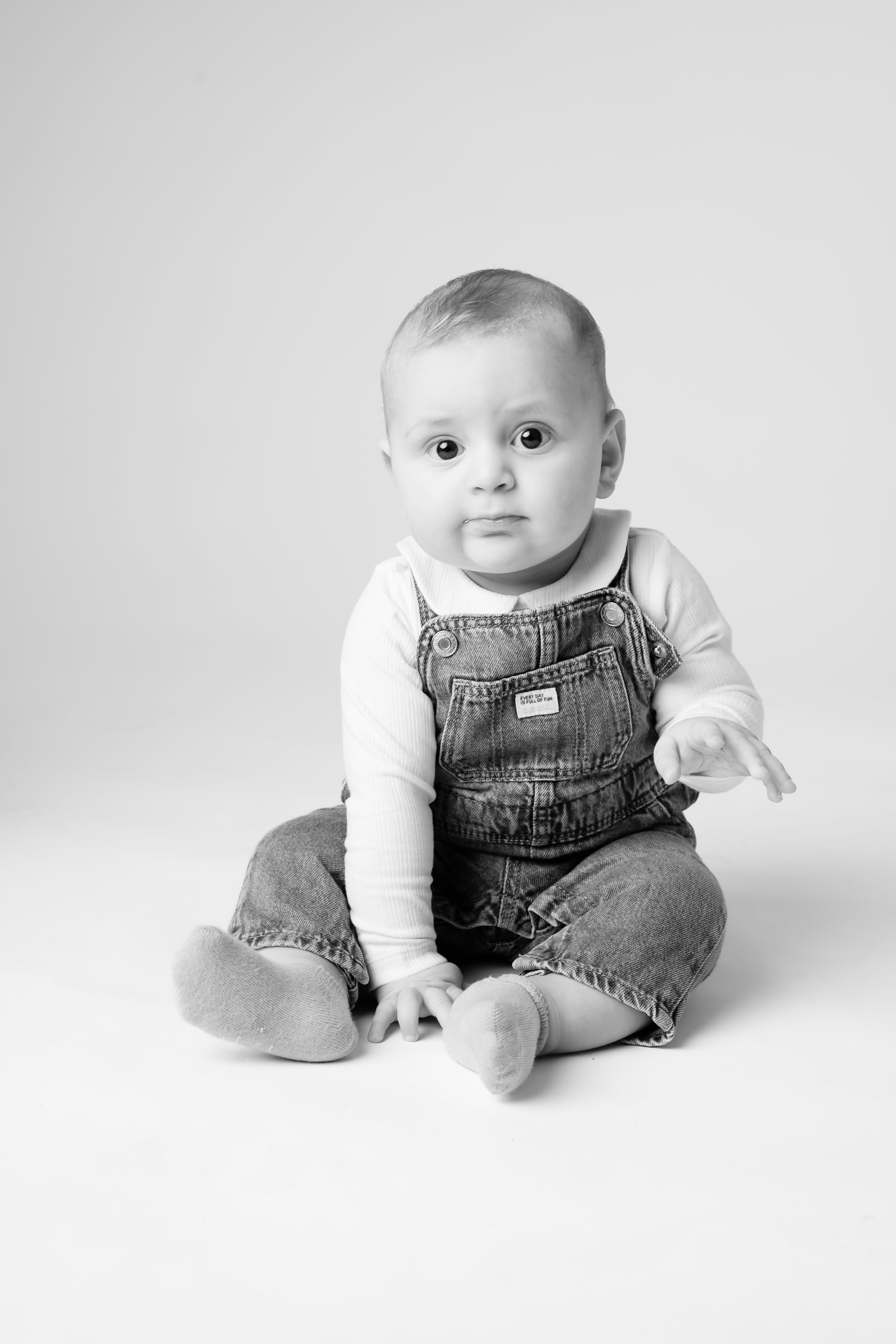 Svart-hvitt-portrett av en baby som sitter på gulvet i skjorte og dongerismekke.