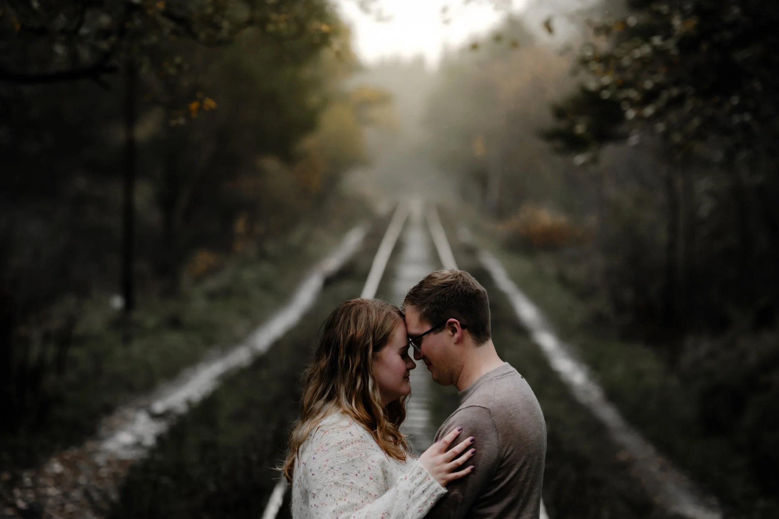 Et par står nært sammen på jernbanespor i skog, holder rundt hverandre og har panna mot hverandre under tåkelagt høstlandskap.