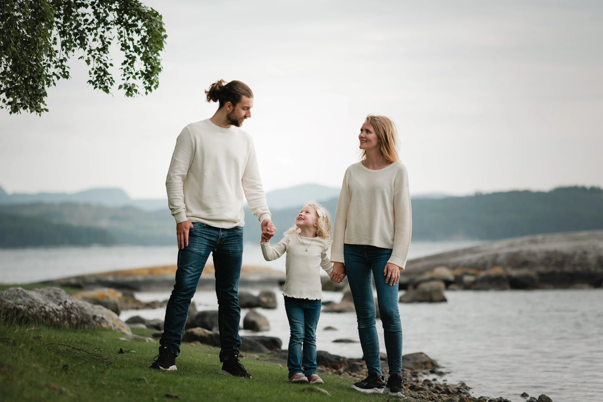 En familie på fire holder hender og går ved en sjø med fjell i bakgrunnen.
