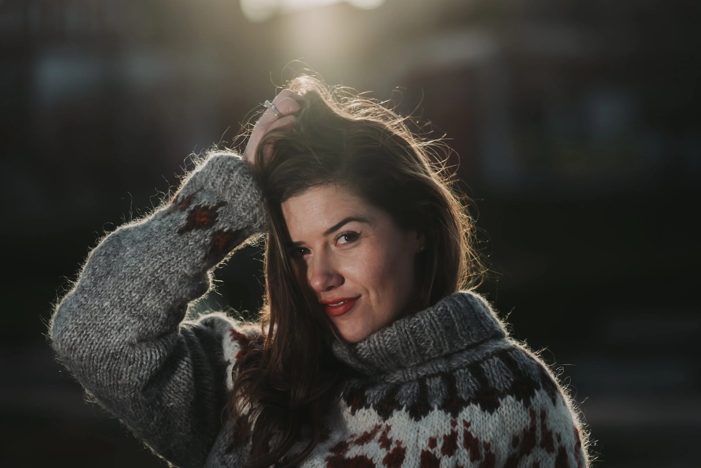 Kvinne med langt brunt hår som smiler, iført en grå og hvit strikkegenser med julemotiv, med hånden i håret, utenfor i solnedgang.
