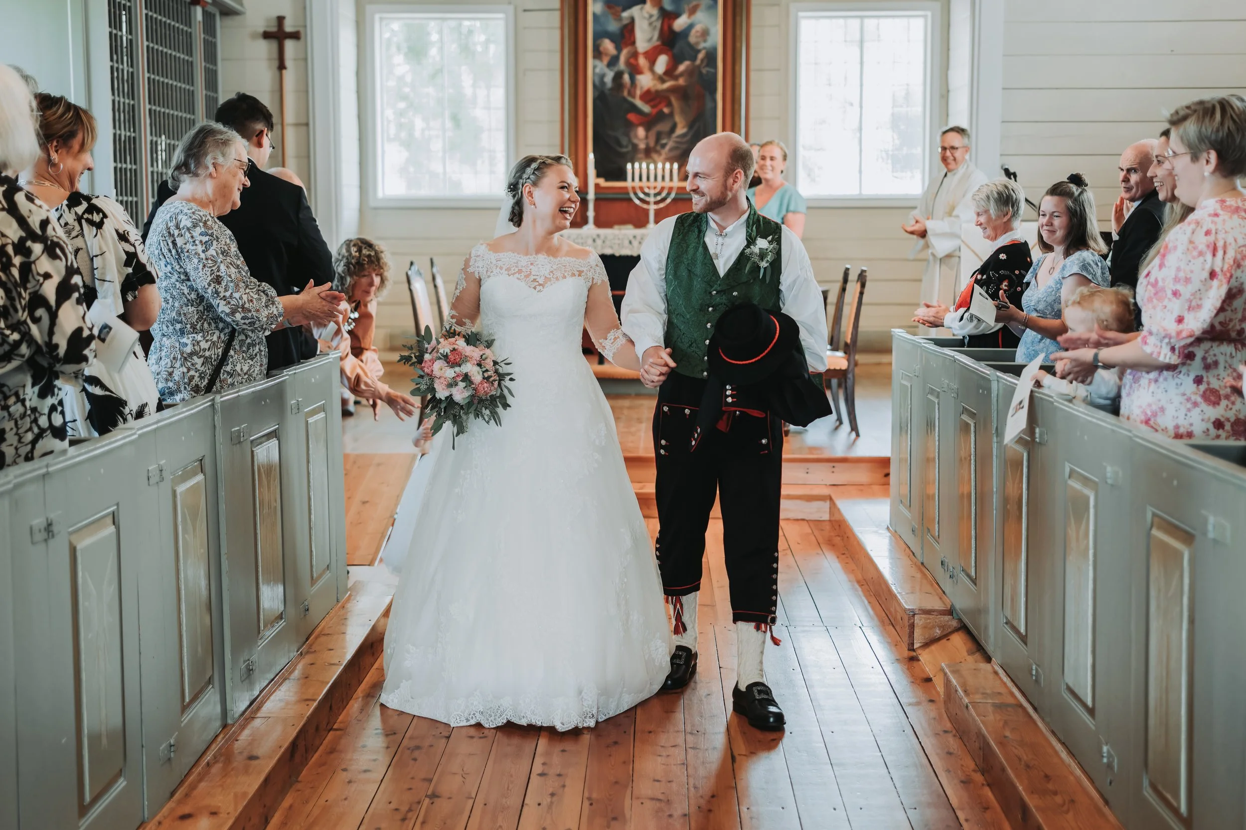 En brud og en brudgom går ned midtgangen i en kirke, omgitt av gjester som smiler og applauderer.