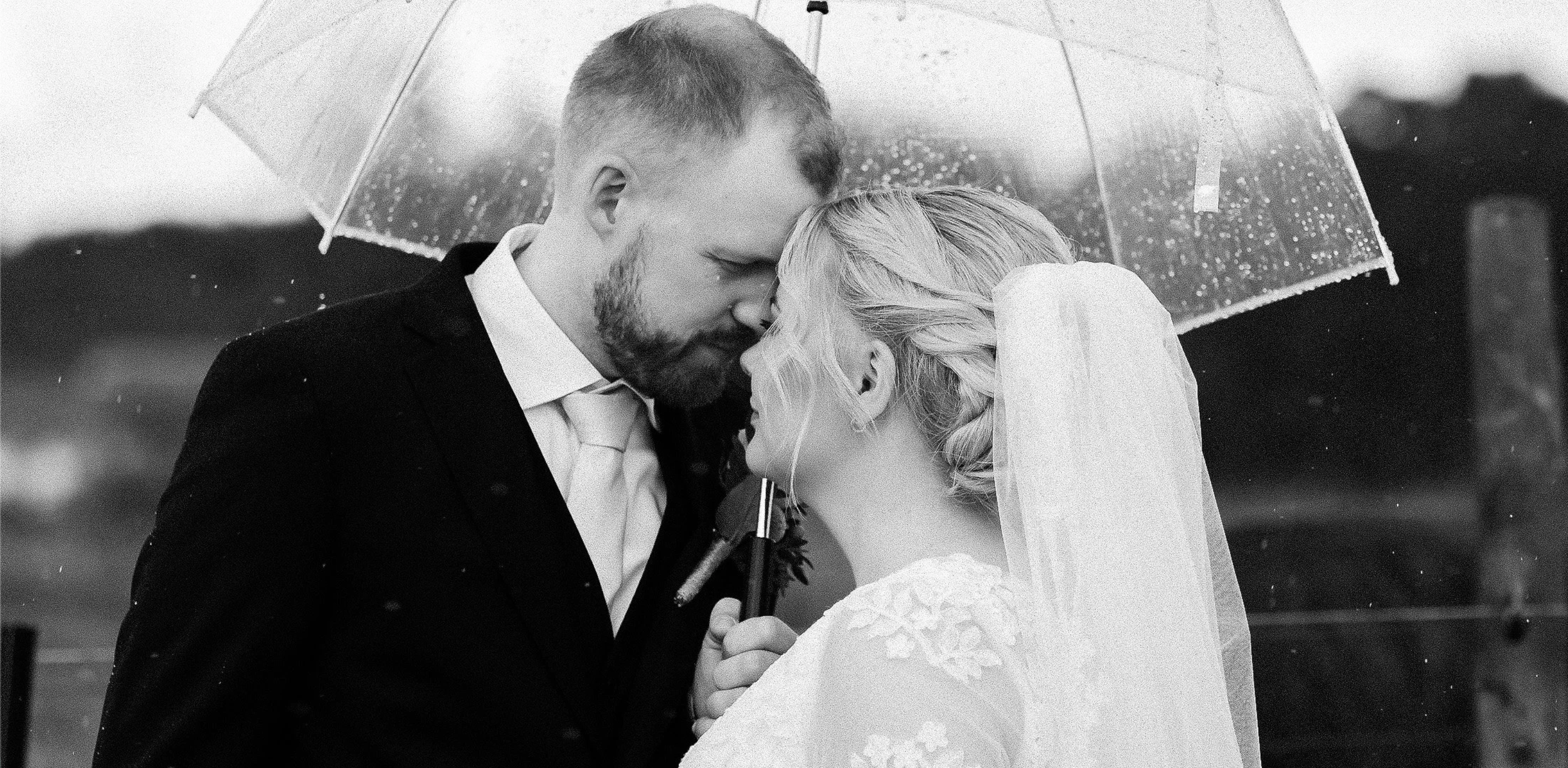 Et brudepar står under en gjennomsiktig paraply i regnet, de kysser med pannen mot hverandre. Det er svart-hvitt bilde.