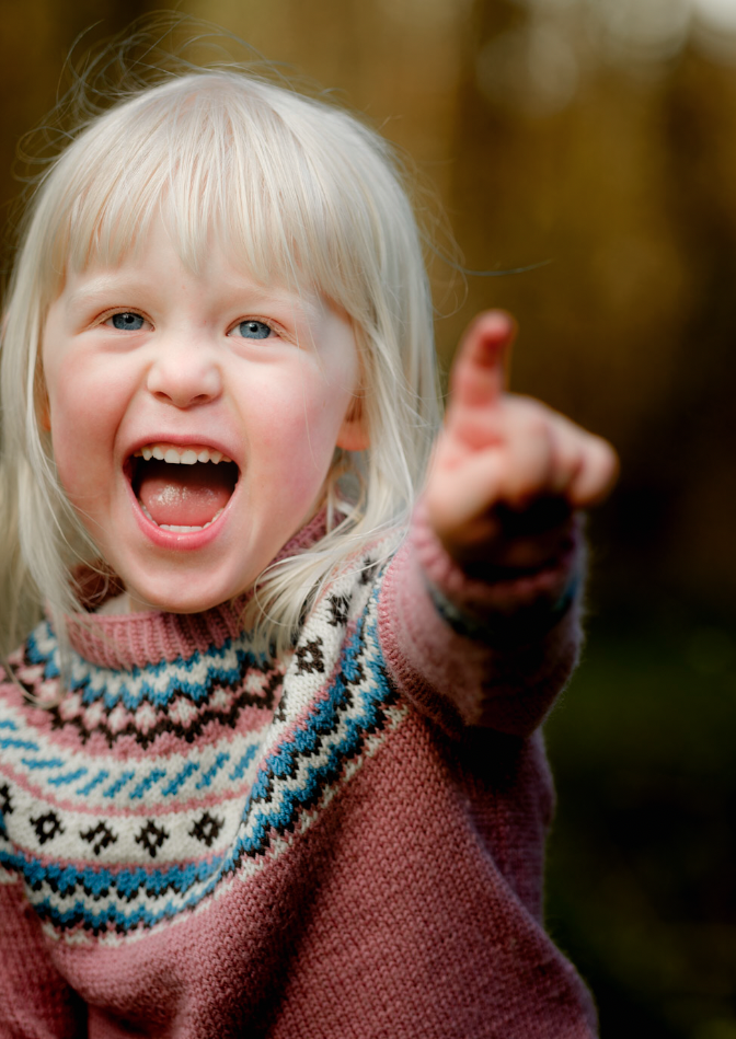 En glad ung jente med blondt hår som ler og peker mot kameraet, utendørs med en bakgrunn av høstfarger.