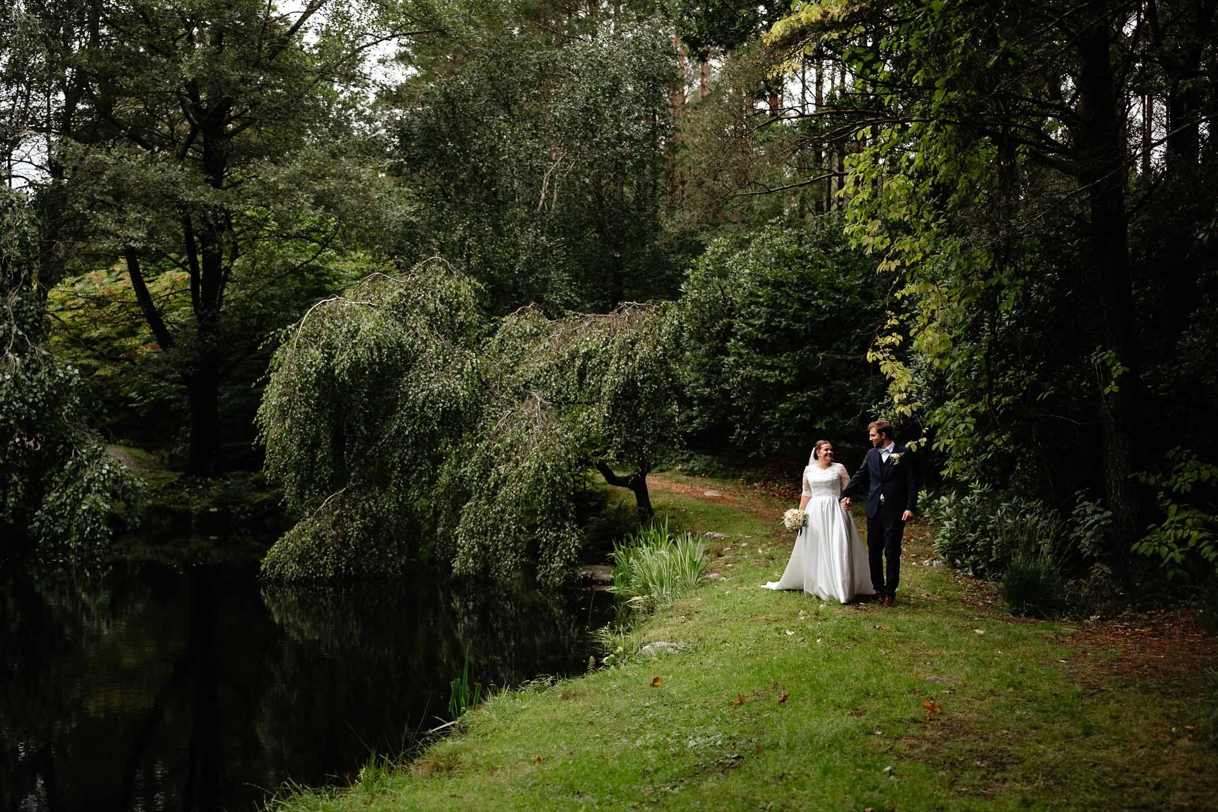 Brudepar går hånd i hånd langs en dam i en frodig skog; bruden bærer hvit kjole og bukett, og brudgommen mørk dress.