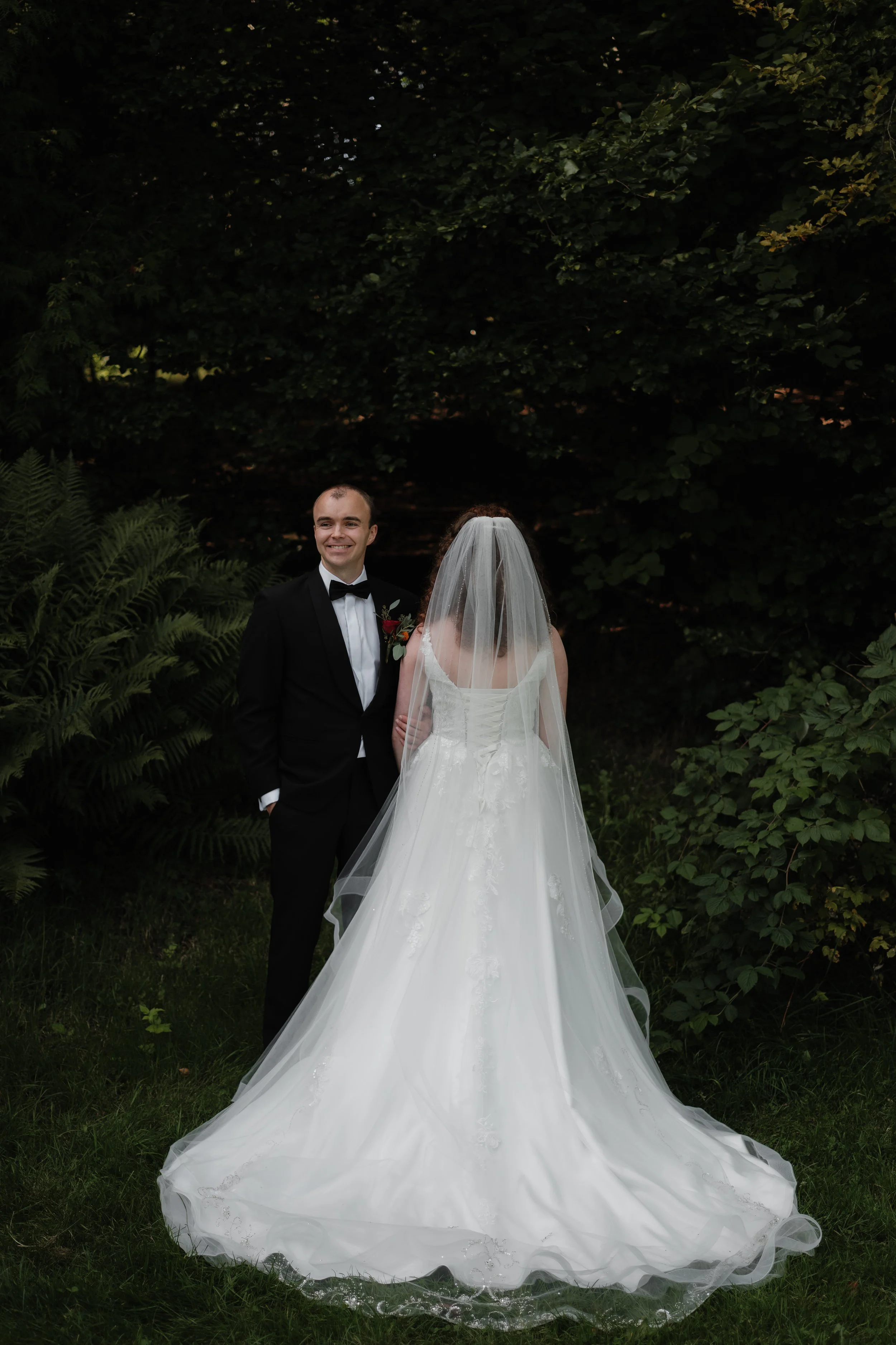 Et brudepar står i en grønn hage, med mannen i en svart smoking med hvit skjorte og sløyfe, og kvinnen i en hvit brudekjole med slør.