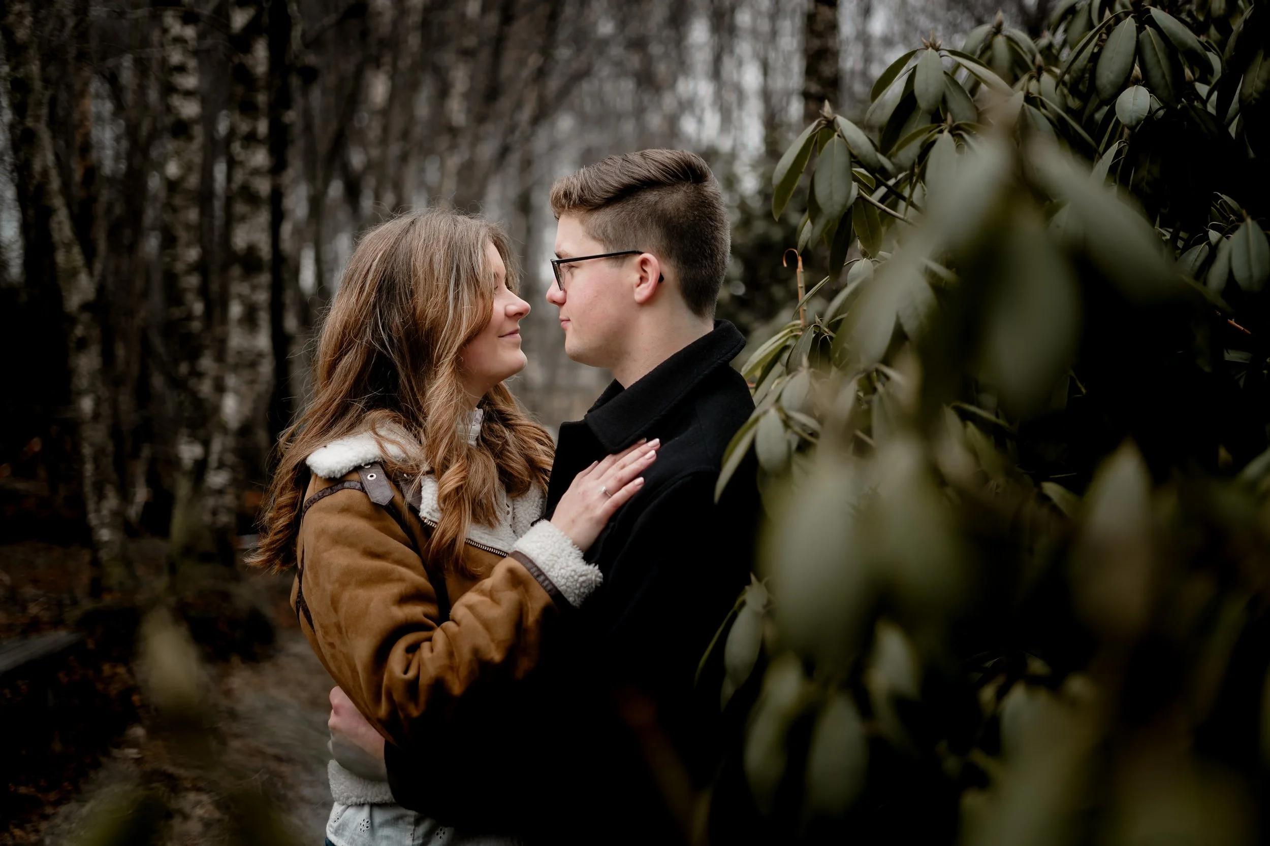 Et par står tett sammen i en skog, ser på hverandre, omgitt av blader og trær, en drømmeaktig scene.