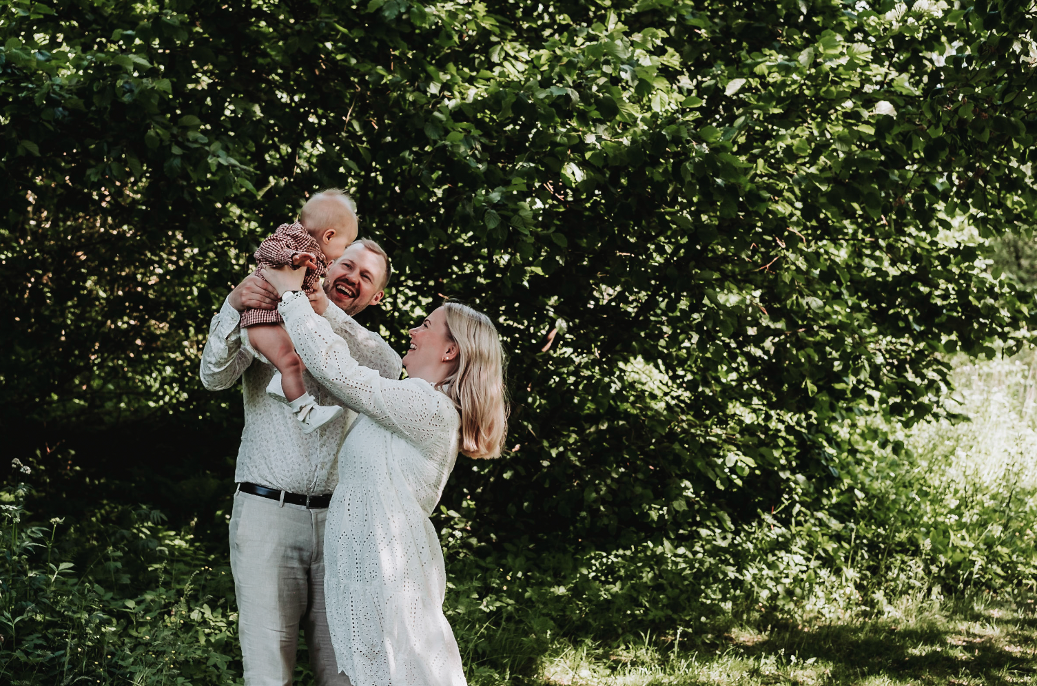 Et lykkelig familie bestående av to voksne og et lite barn, som smiler og leker ute under trær.
