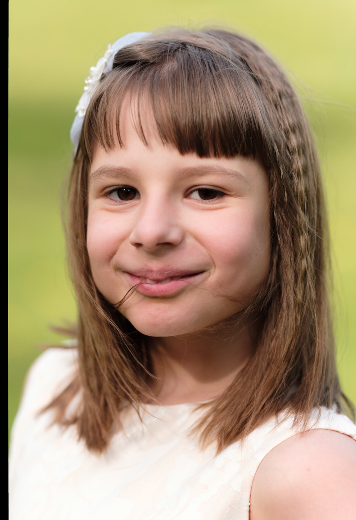 Et portrett av en ung jente med brunt hår og humørfylt ansiktsuttrykk, iført en hvit kjole, utendørs med grønn bakgrunn.