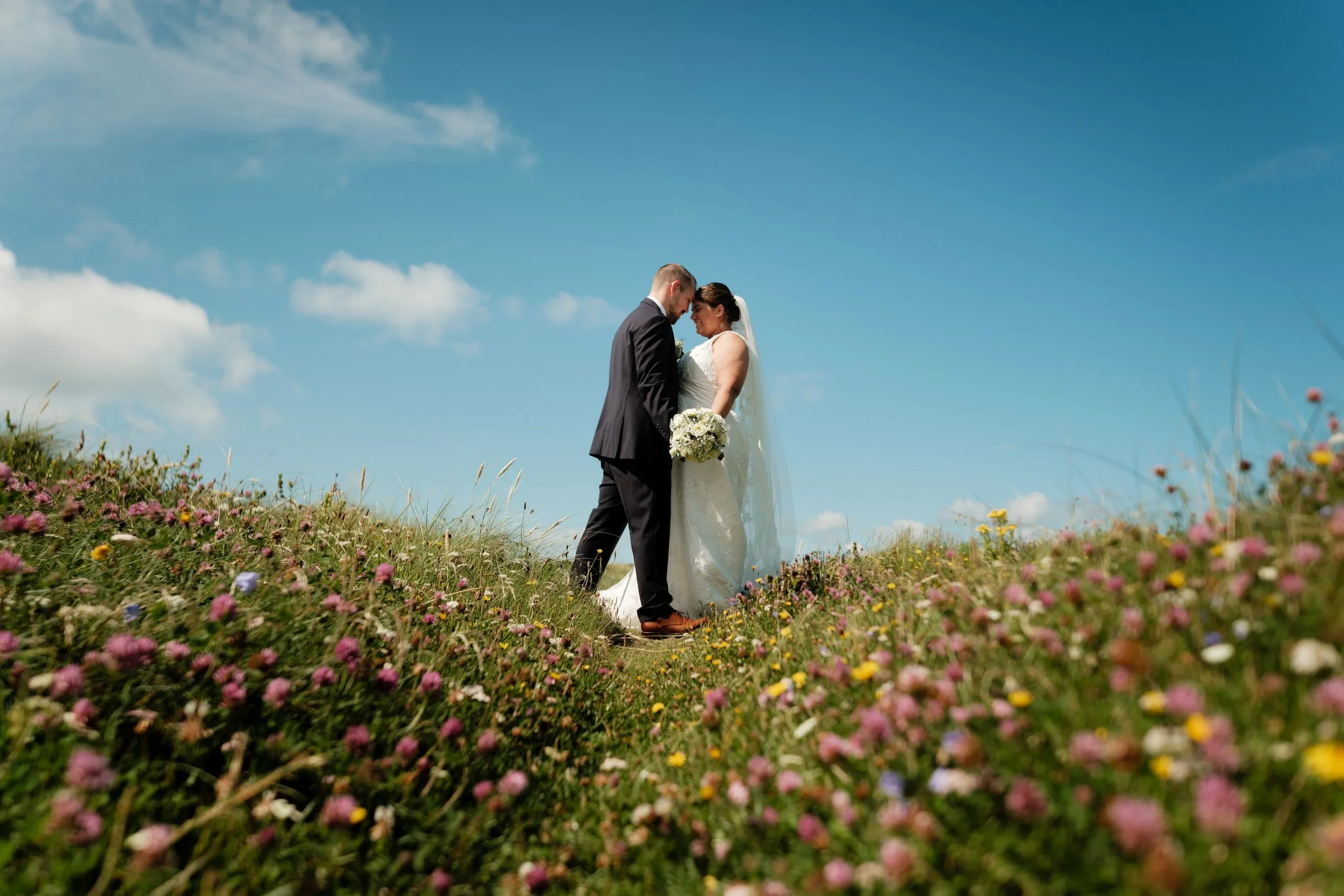 Brudepar omfavner hverandre i en blomstereng under en blå himmel