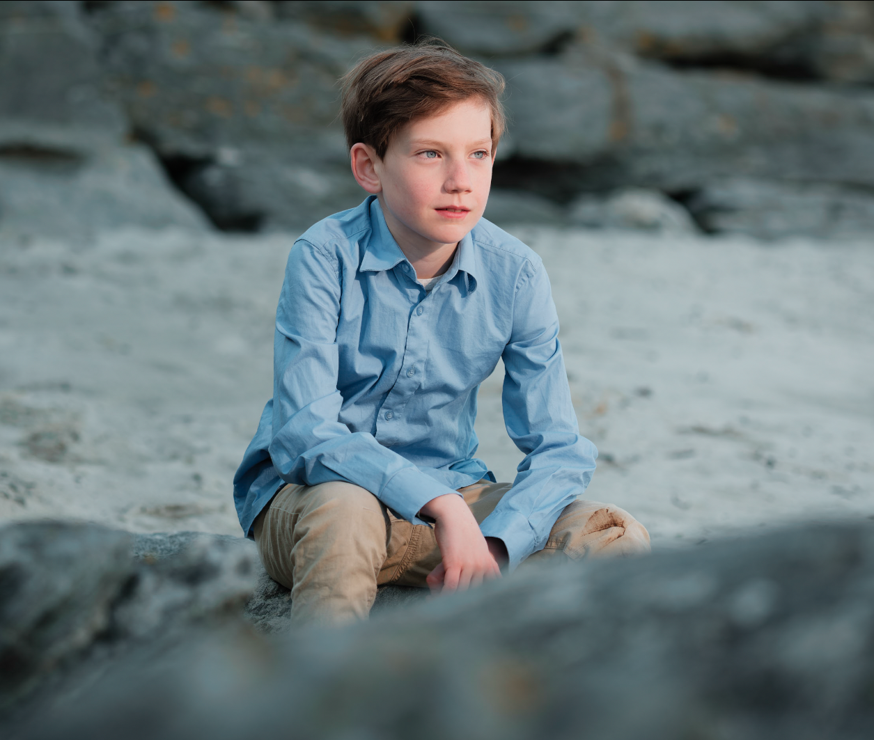 En unge med blå skjorte og beige bukser sitter på en stein ved stranden og ser ut i det fjerne.