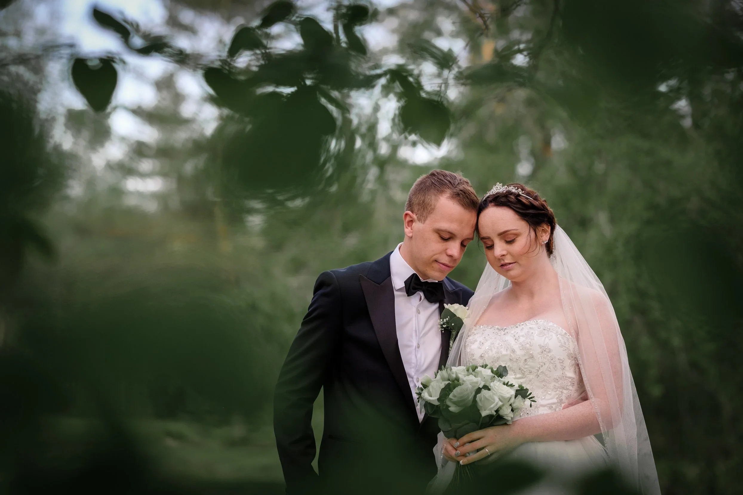 Et bryllupsbilde av et ektepar i brudekjole og smoking, som står under trær og holder en blomsterbukett, med lav dybdeskarphet og naturlig utendørs bakgrunn.