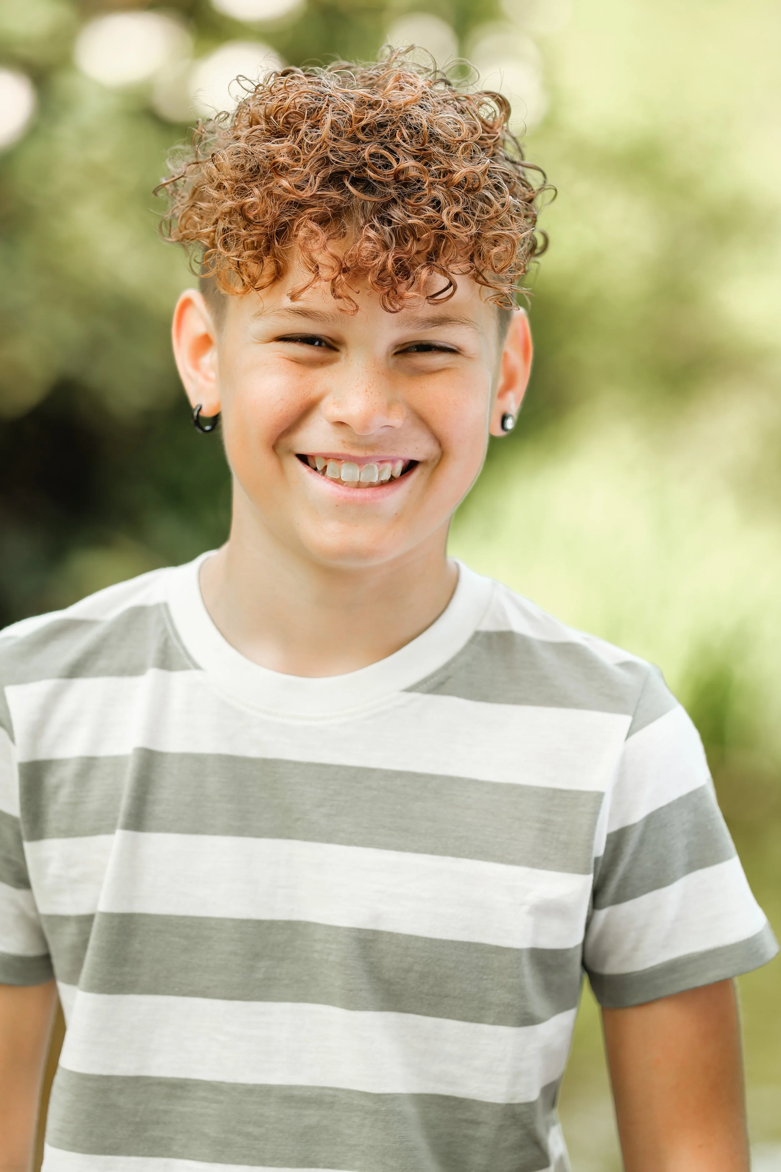 Smilende gutt med krøllete, lyst brunt hår og hull i ørene, iført en stripet t-skjorte, utendørs med grønne, uklare bakgrunnsurtsetning.