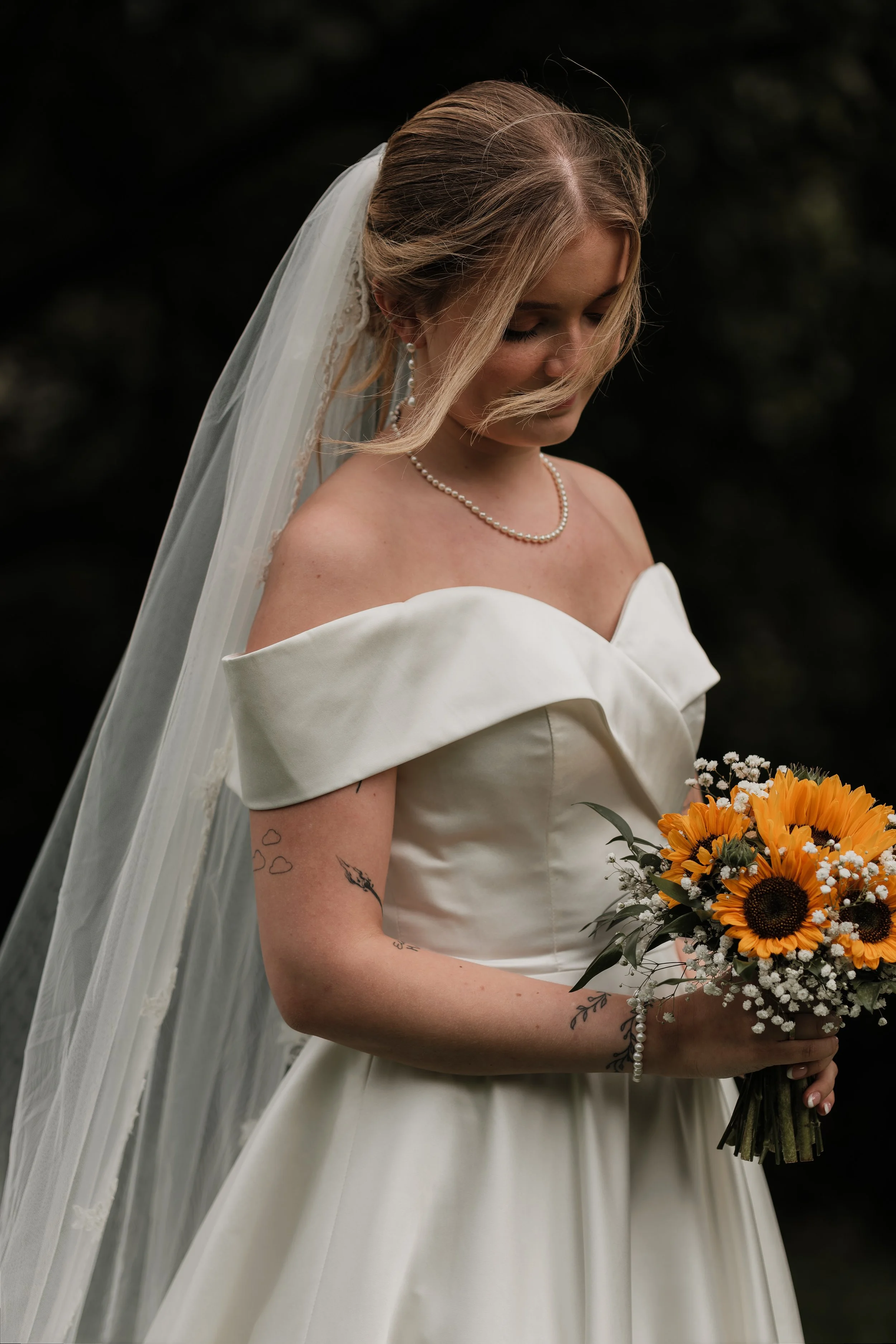 En brud i off-shoulder brudekjole holder en blomsterbukett med solskinnsblomster og hvite små blomster.