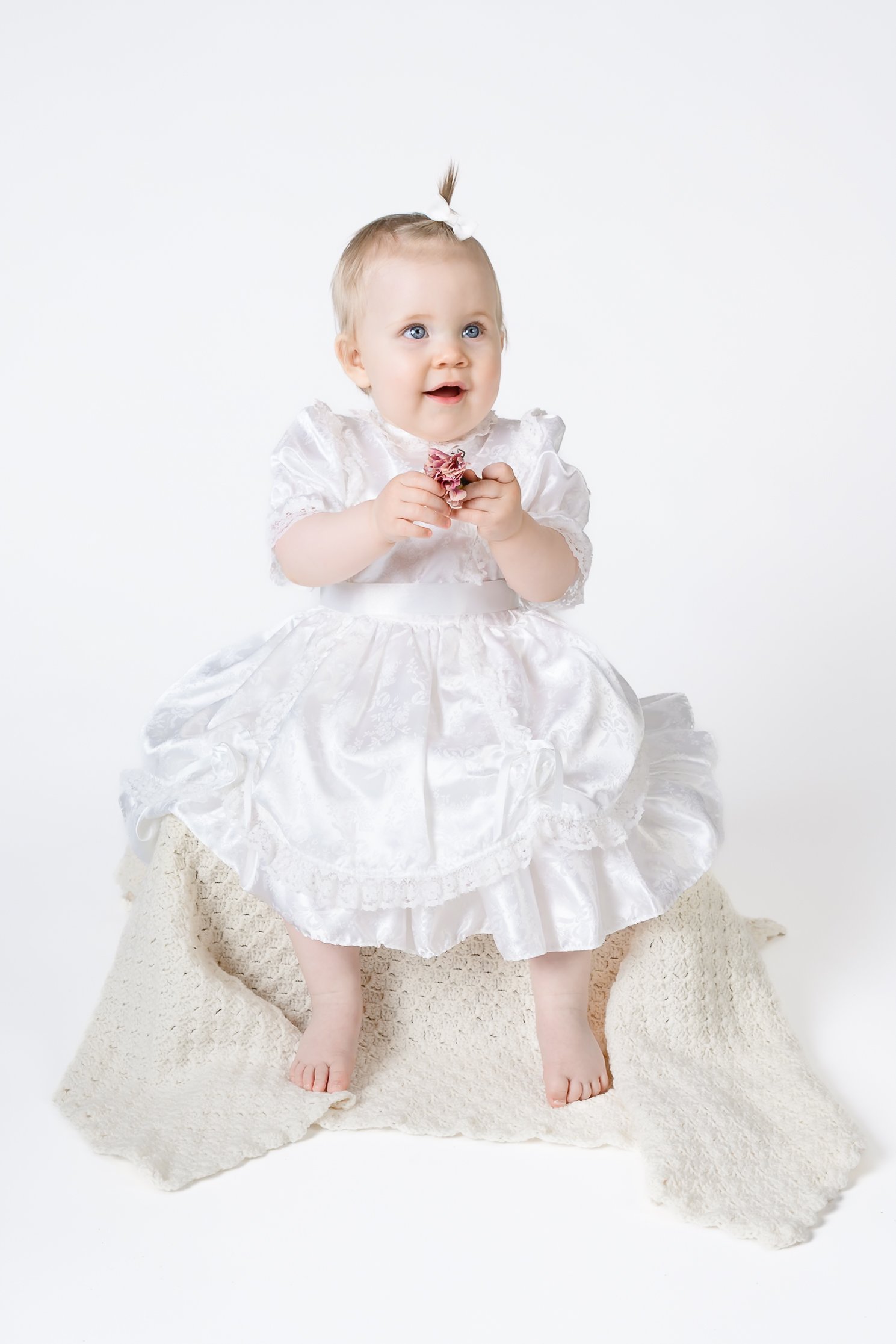 Baby i hvit kjole med glitrende detaljer, som sitter på en beige saueskinn på gulvet, og holder en liten blomsterbukett. Sitter på et hvitt bakgrunn