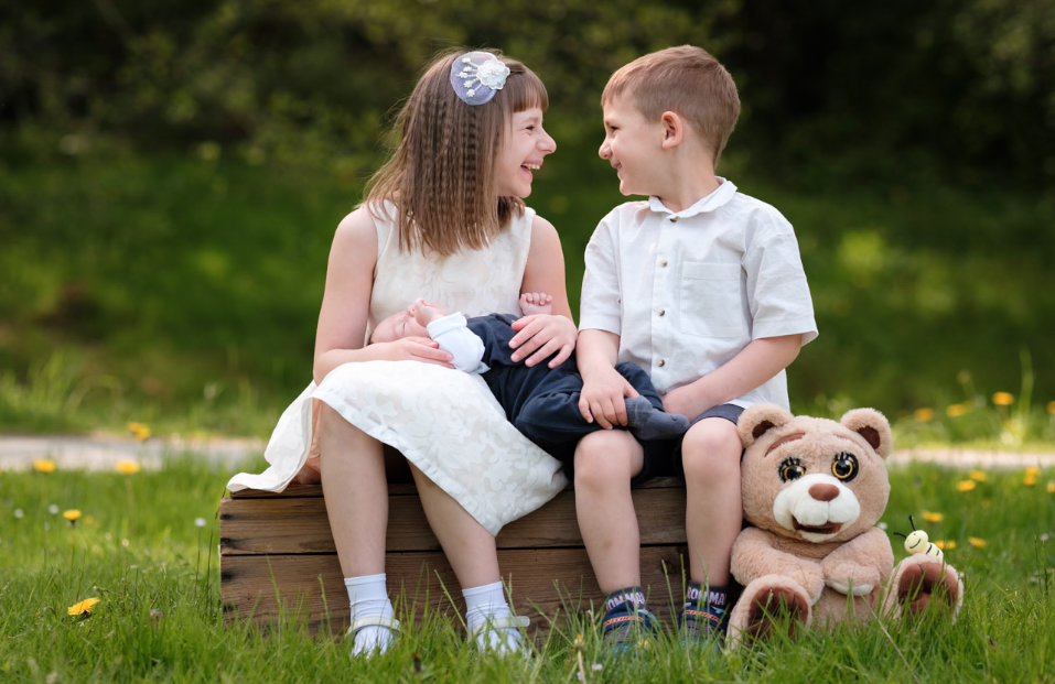 To barn, en jente og en gutt, sitter på en trebenk i en grønn park med blomster, smiler og ser på hverandre. Jenta holder en liten baby, og det er en stor leketøys-isenkatt ved siden av dem.