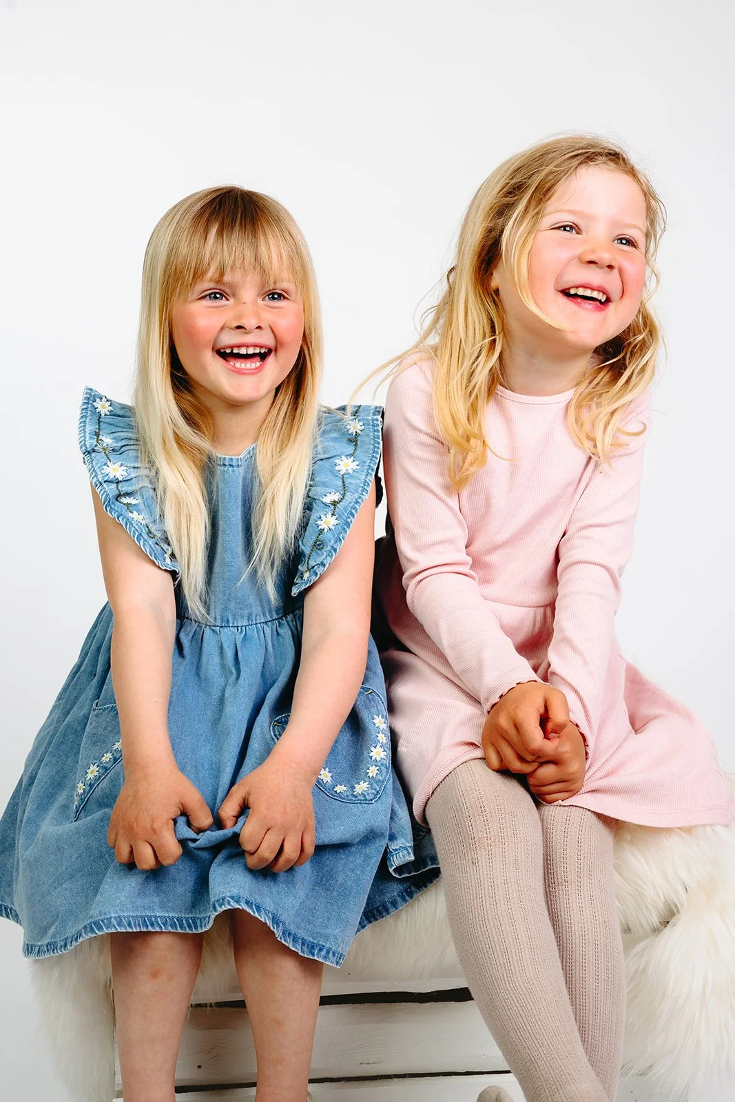 To smilende jenter sitter sammen; den ene har blå denimkjole med blomster, den andre rosa kjole og strømpebukser.