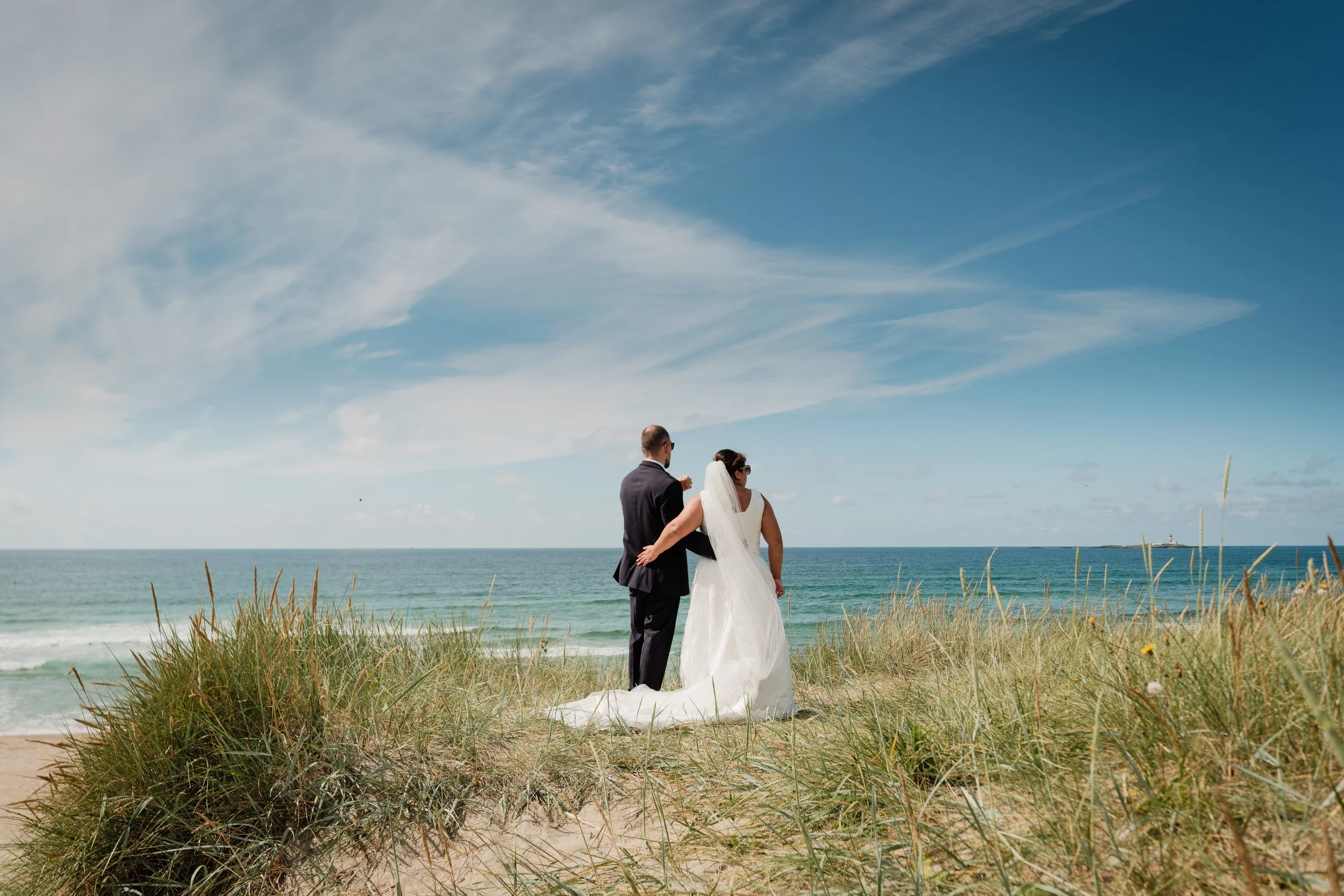 Et brudepar står på en sandstrand med gress, ser utover havet under en blå himmel med noen skyer.