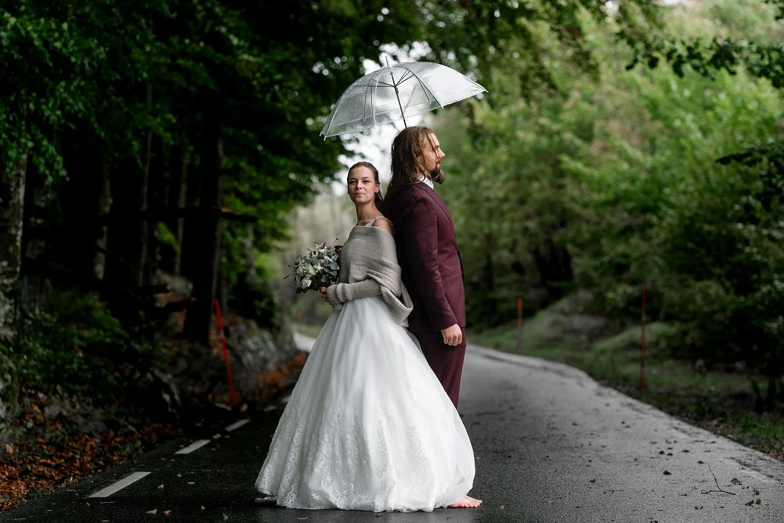 Et brudepar står på en våt vei i en grønn skog, med en kvinne i en hvit brudekjole som holder en bukett blomster og en mann i en mørk dress under en gjennomsiktig paraply.