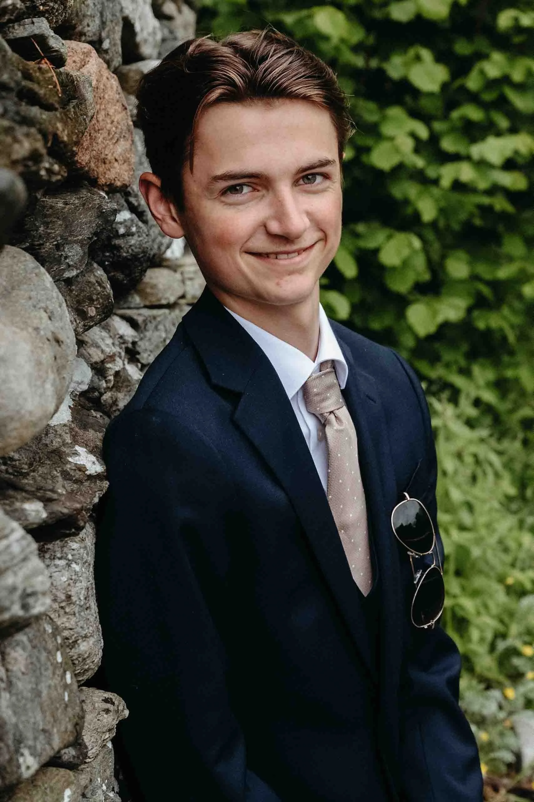 Portrett av en smilende ung mann i mørk dress og slips, stående foran en steinmur og grønt løvverk.