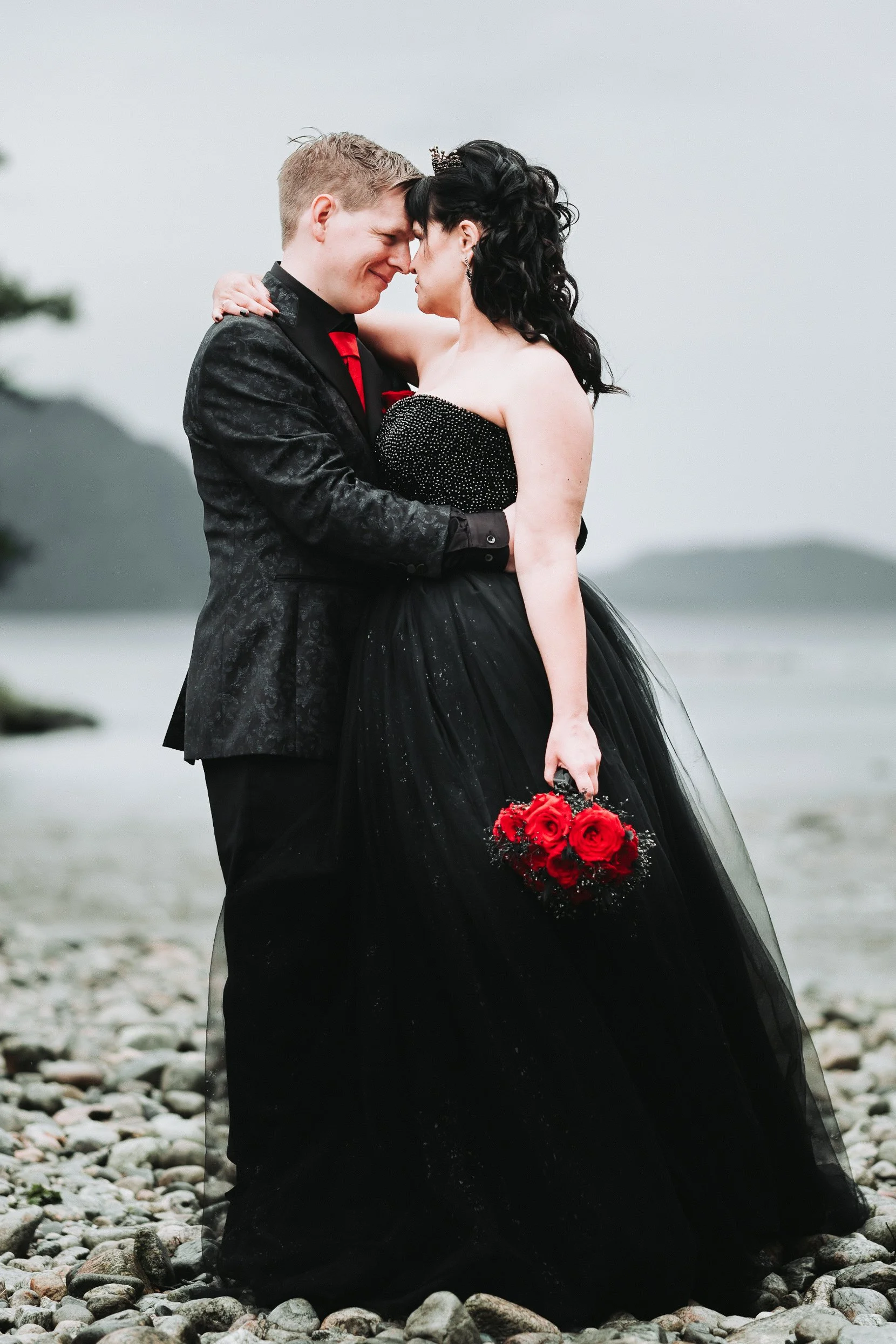 Et bryllupspar som holder hverandre tett på en steinstrand, med fjell og vann i bakgrunnen, kledd i formelle klær, kvinnen holder en bukett med røde roser.