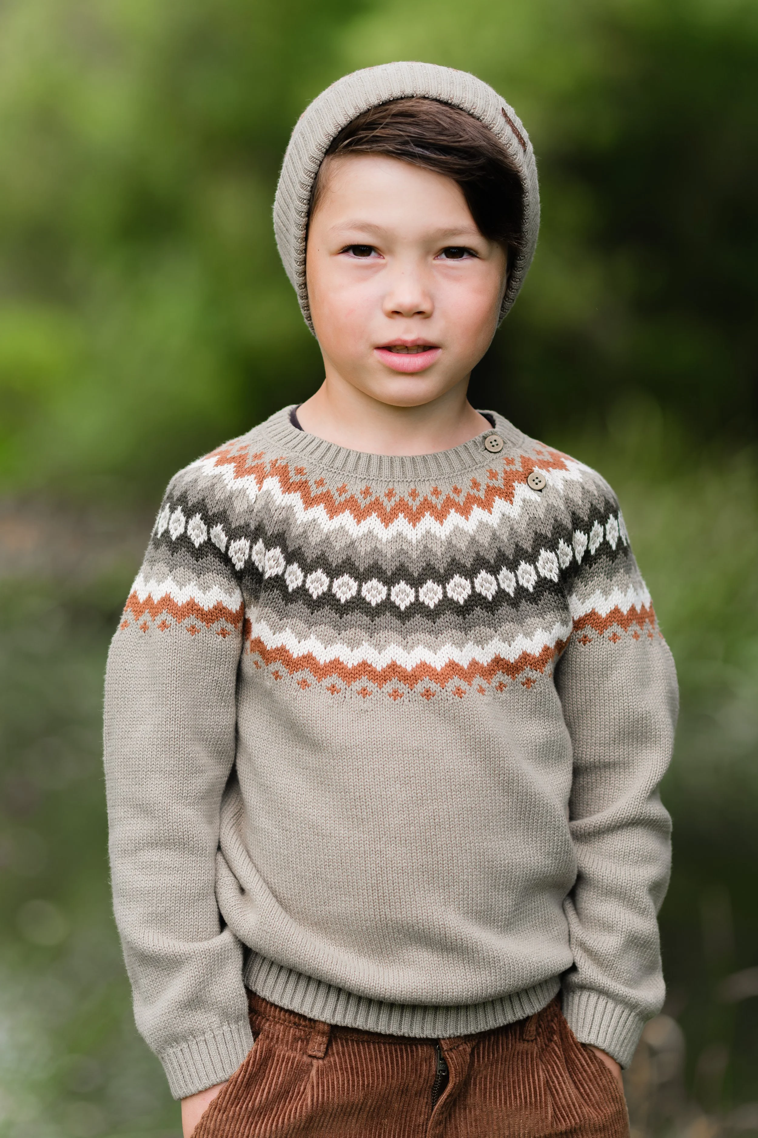 ung gutt som står utendørs i en park med grønt løv bakgrunn, iført en beige genser med mønster i brunt, hvitt og grått, og en grå lue