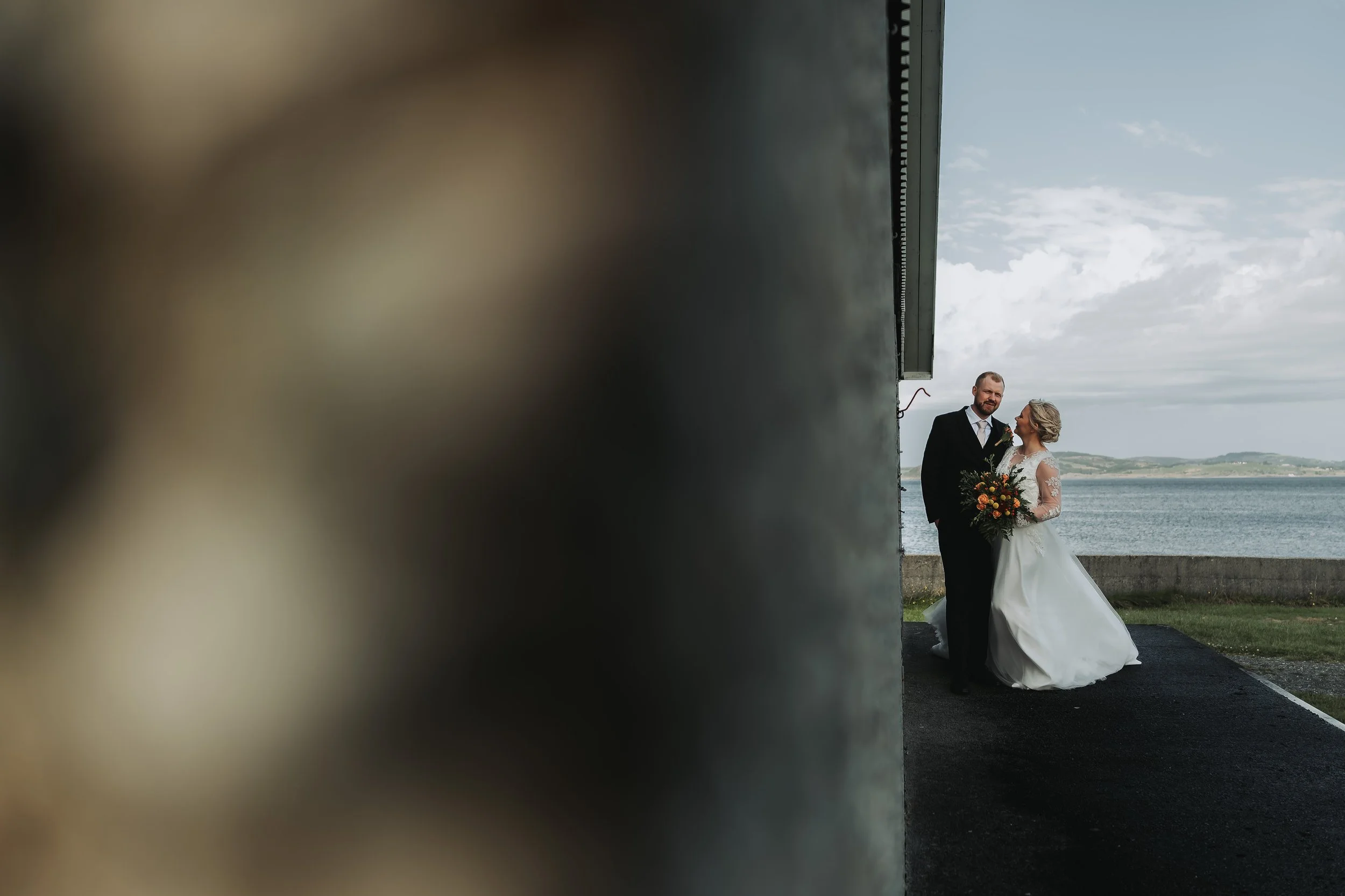 Bryllupsbilde av et par, en mann i dress og en kvinne i brudekjole, fotografert utendørs ved sjøen med en grå murvegg i forgrunnen delvis ut av fokus.