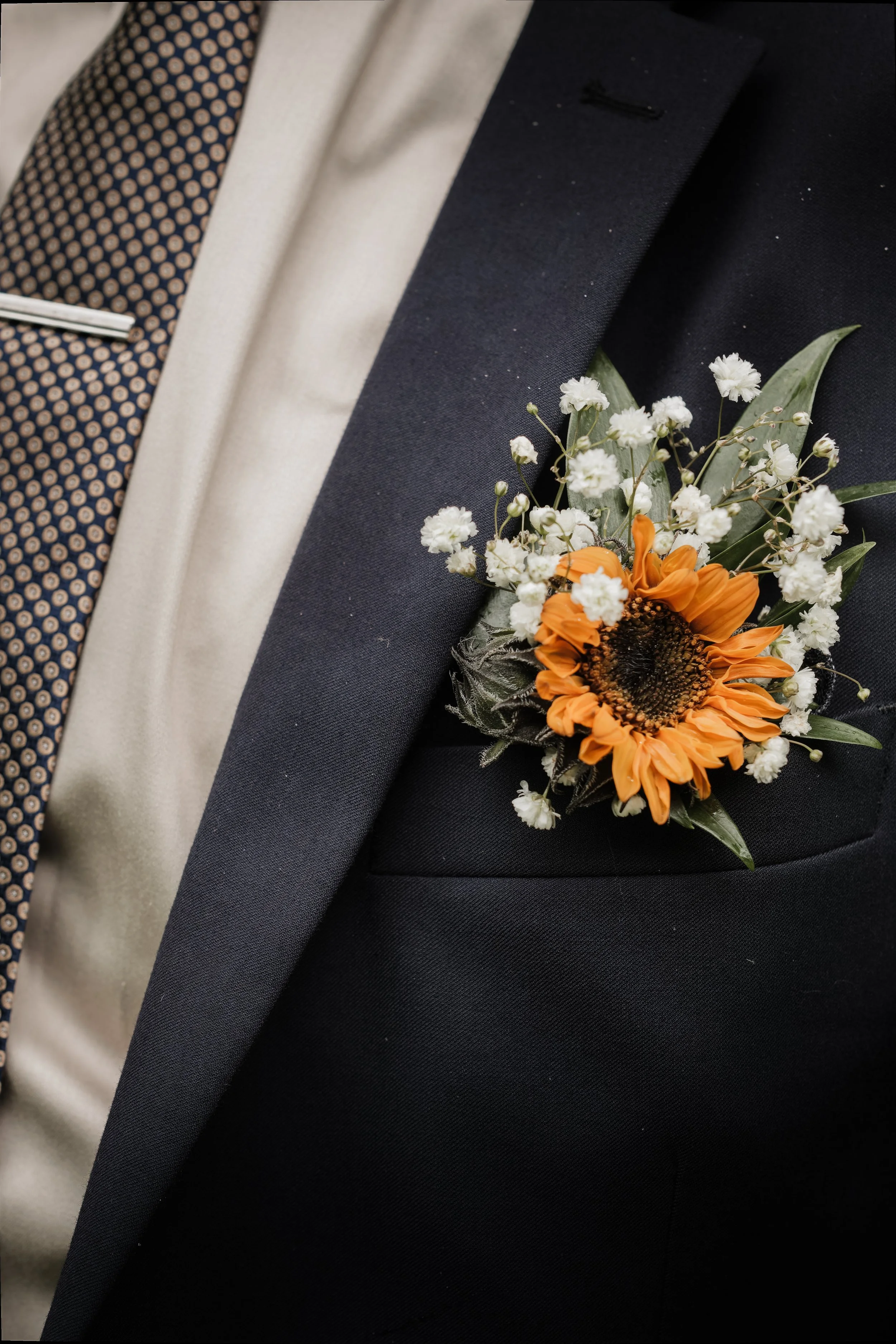 En mann i dress, med hvit skjorte og slips med prikkemønster, har en blomsterlapp med en solrose, småhvite blomster og grønne blader i brystlomma.