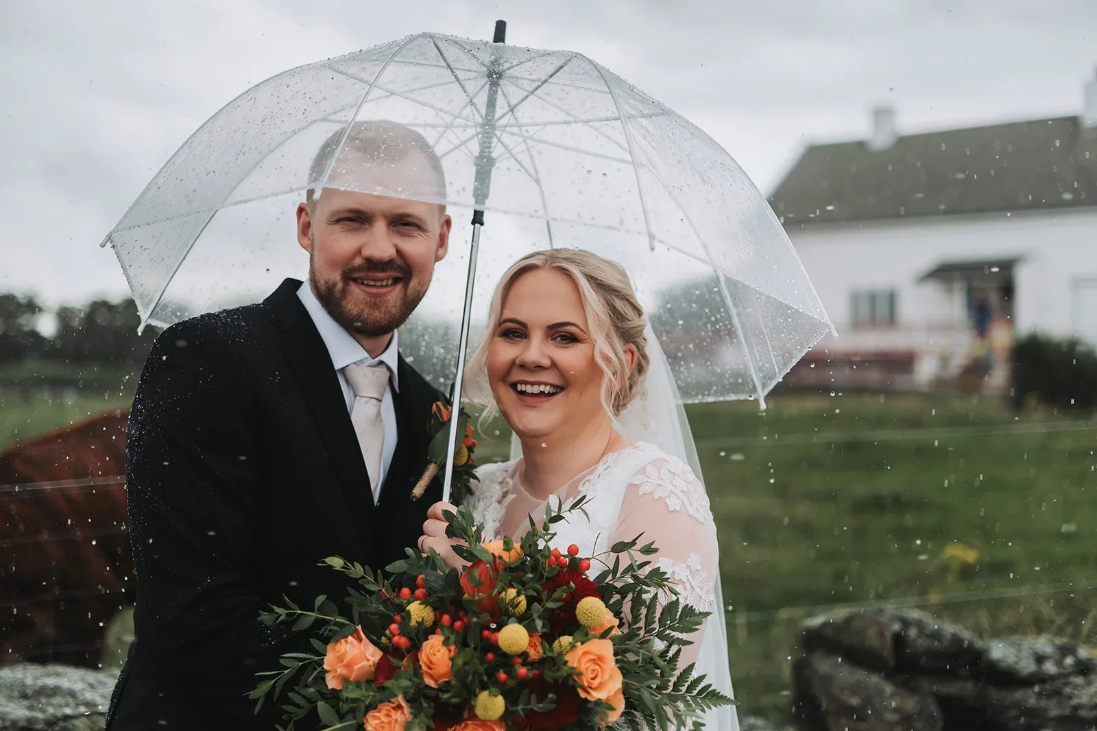 Brudepar smiler under en gjennomsiktig paraply i regnvær, med brudebukett foran et hus.