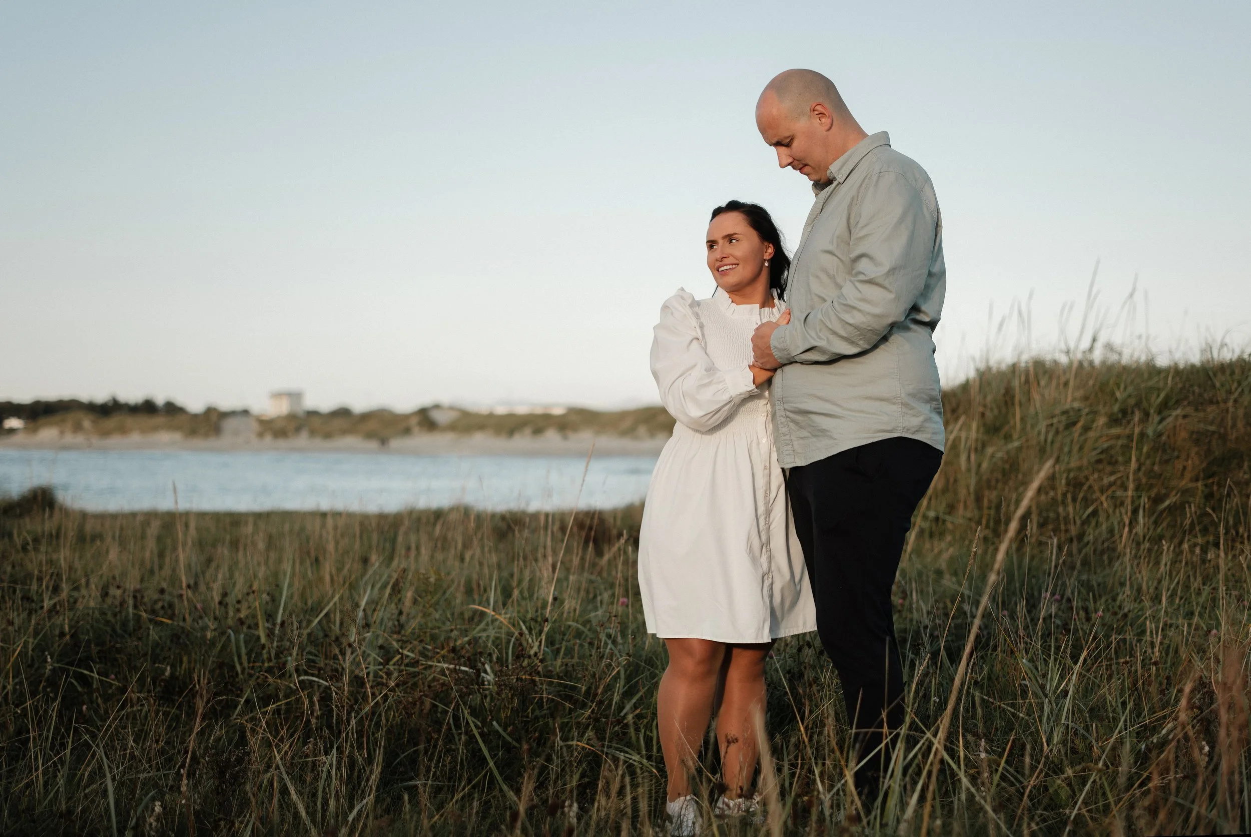 Et par står i naturen nær en innsjø om kvelden, omfavner hverandre og smiler.