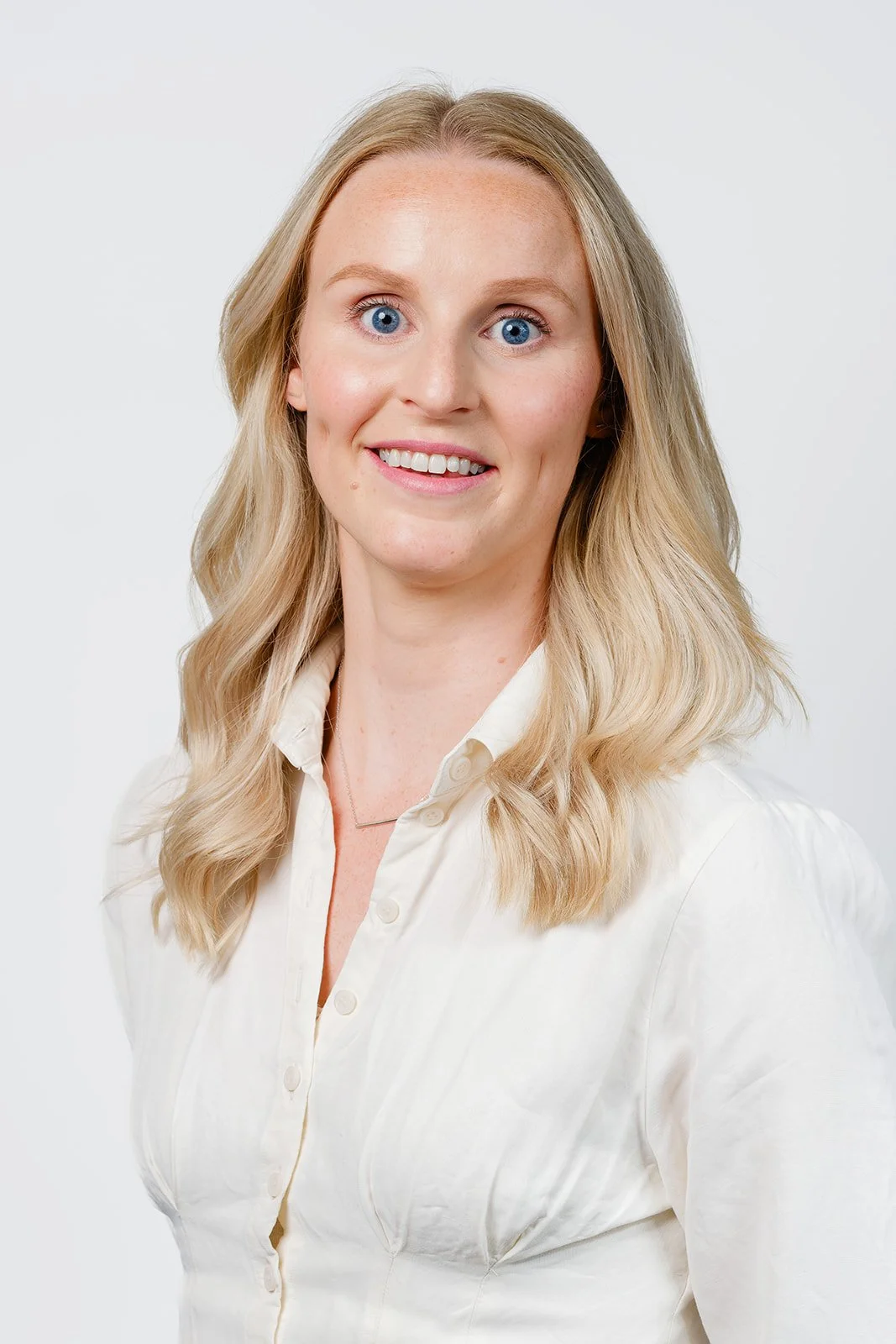 Smilende blond kvinne med blå øyne i hvit skjorte mot lys bakgrunn