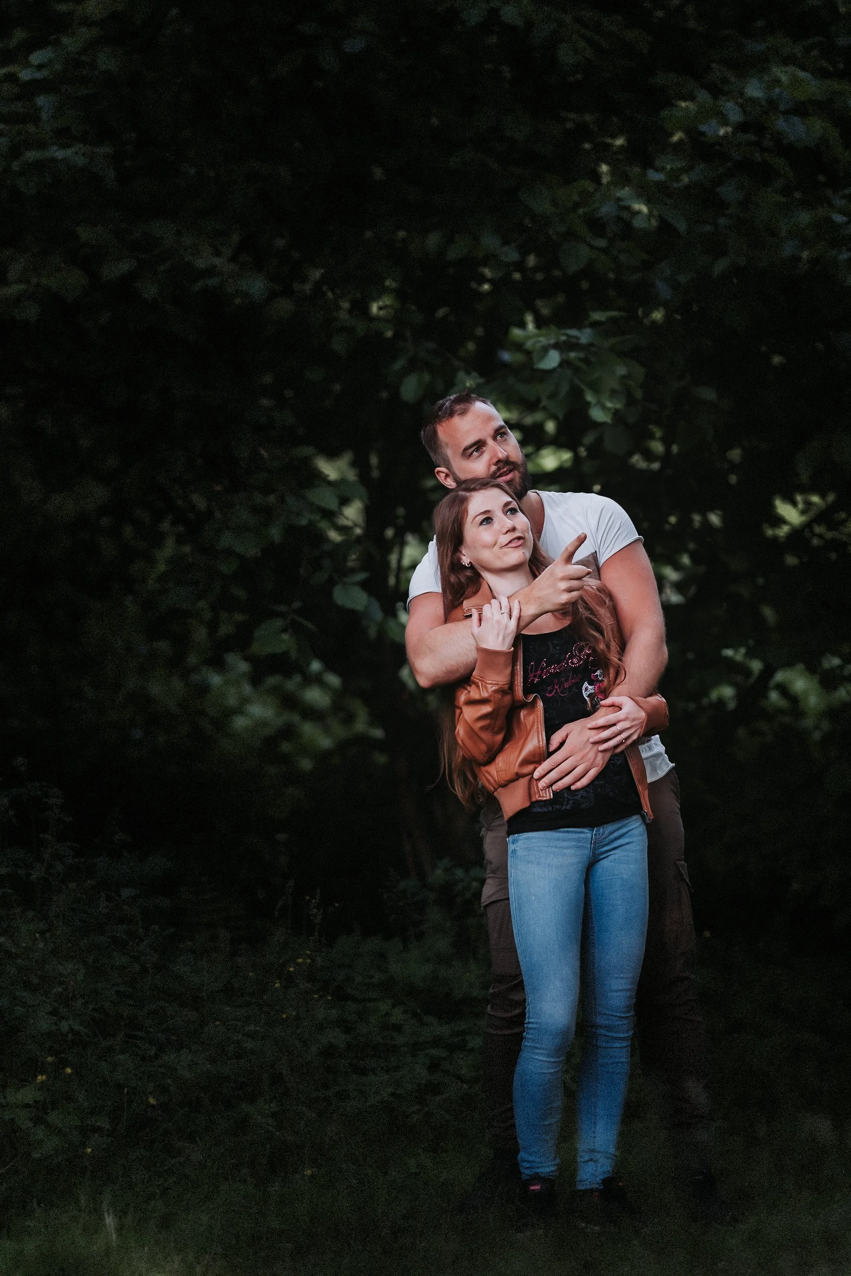 Et ungt par og en ung kvinne står sammen i en skog, den unge mannen holder rundt kvinnen og viser noe med hånden, begge ser oppmerksomme ut.