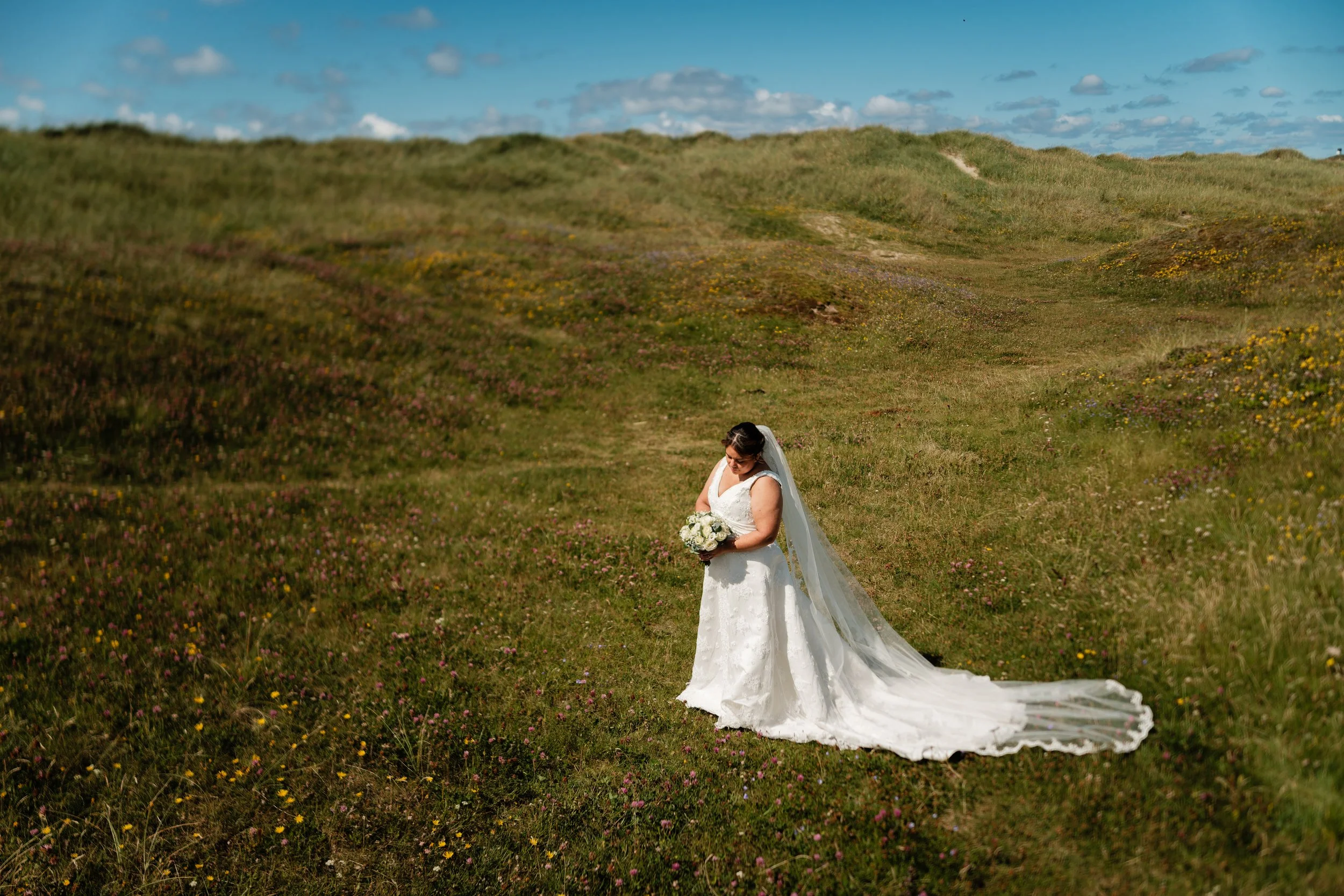 En brud i hvit kjole og slør som står i en grønn åslandskap og holder en blomsterbukett.