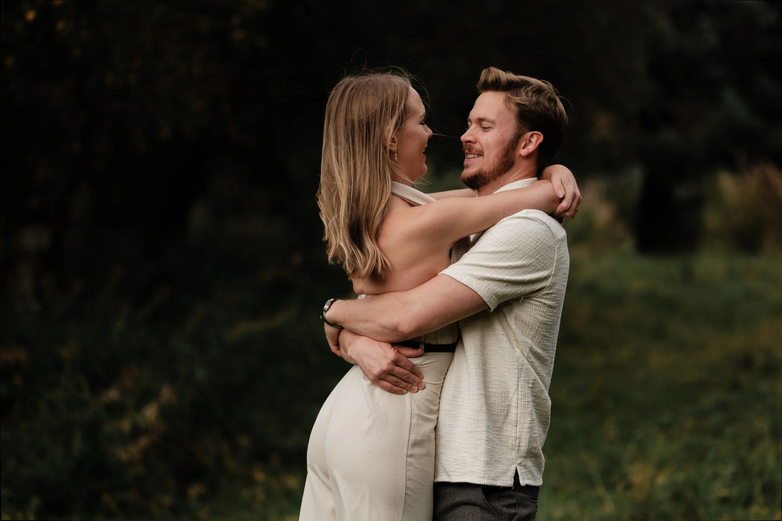 Et par omfavner hverandre i en utendørs park, smilende og glade.