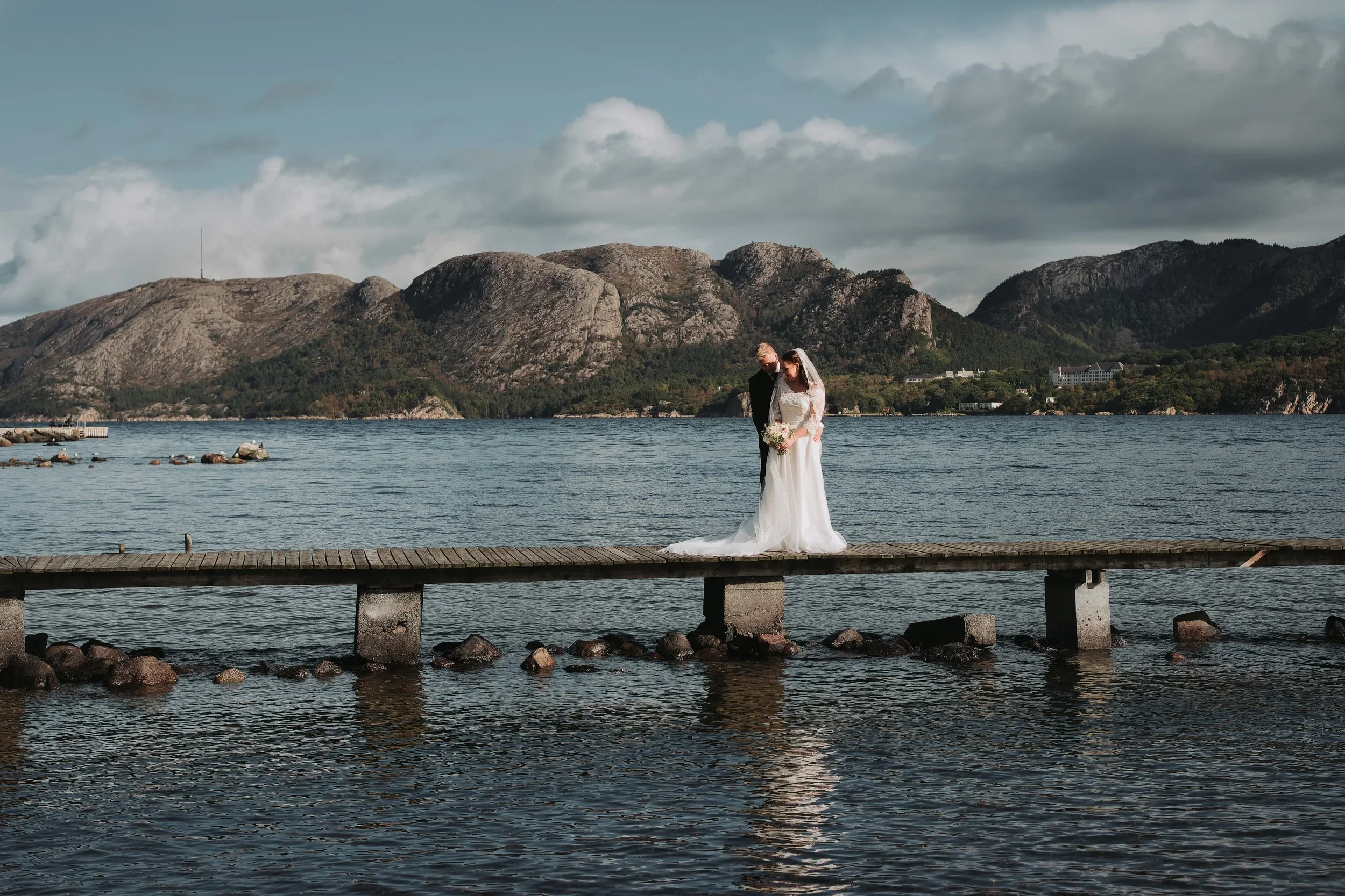Brudepar på en trebrygge over vannet, med fjellandskap og overskyet himmel i bakgrunnen.