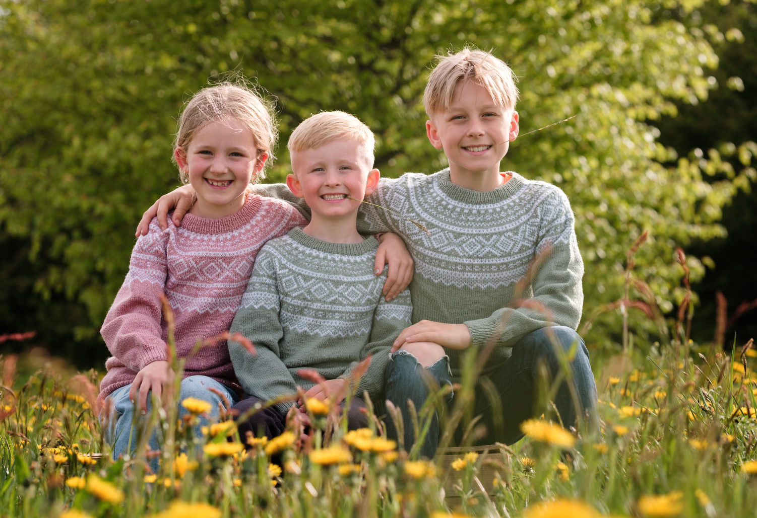 Tre barn som smiler og sitter i en blomstereng på våren, med grønne trær i bakgrunnen, alle iført strikkede gensere.