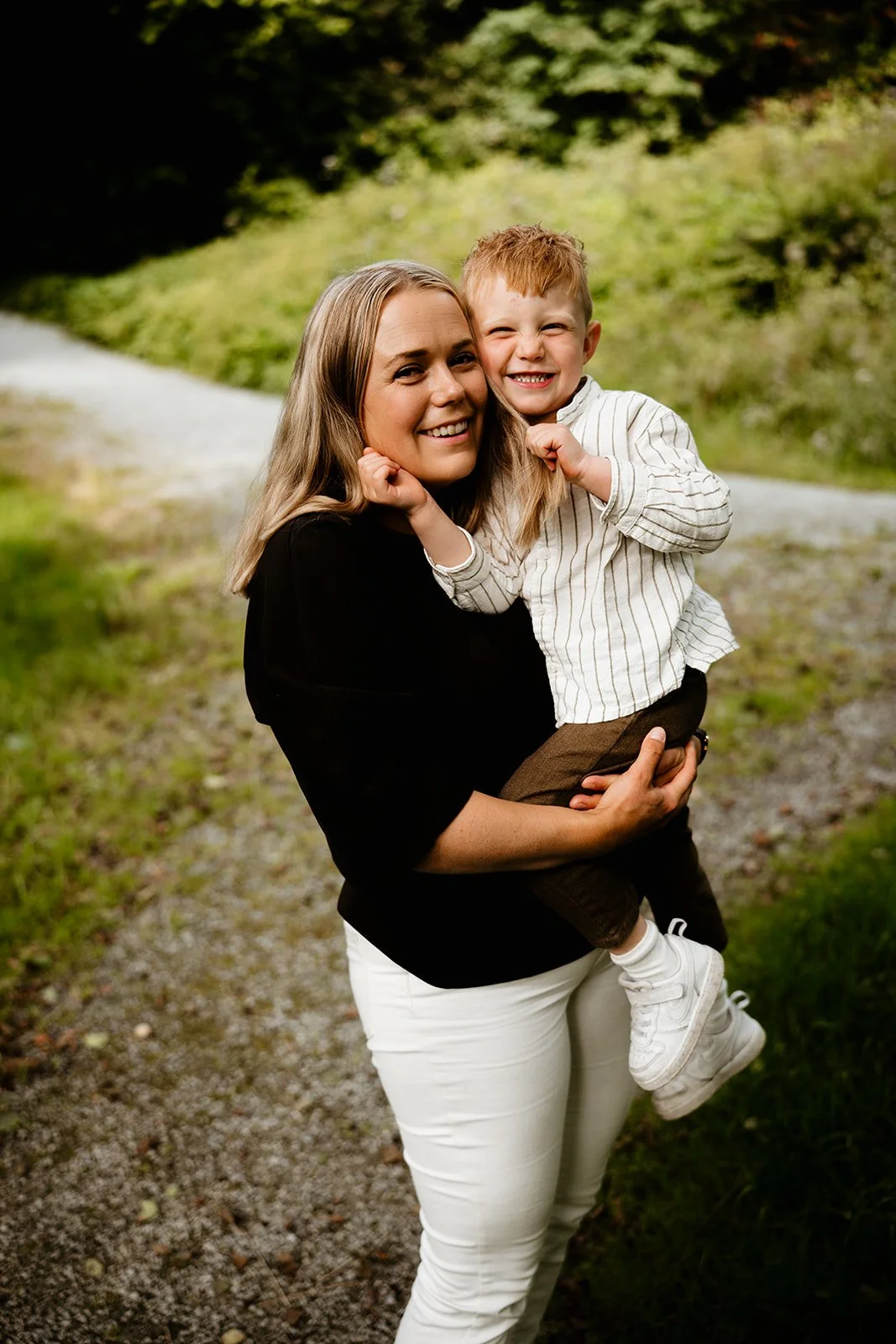 Smilende mor holder en liten gutt på armen utendørs ved en grusvei.