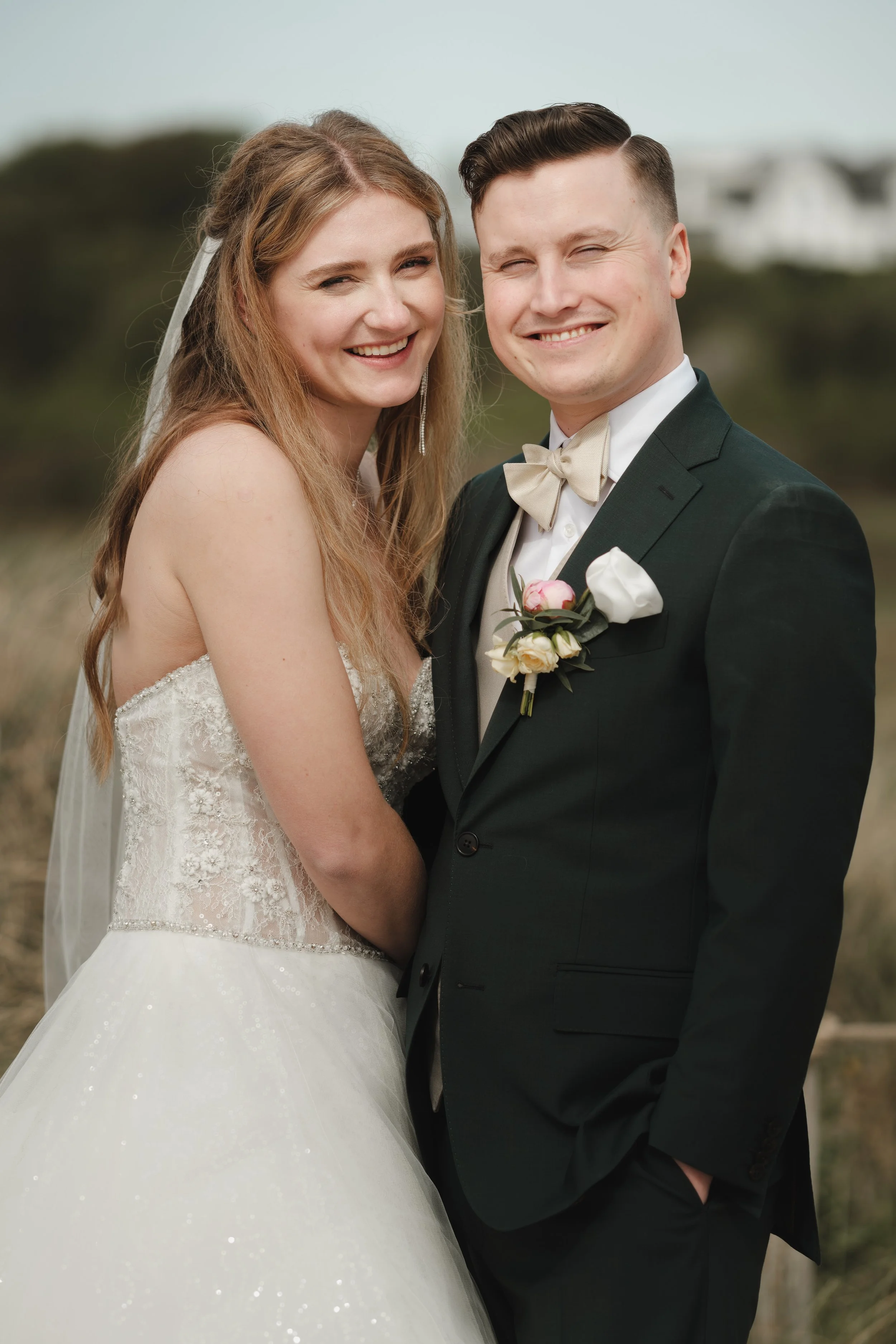 Et brudepar står ute, kvinnen i brudekjole og mannen i dress med boutonnière, smilende