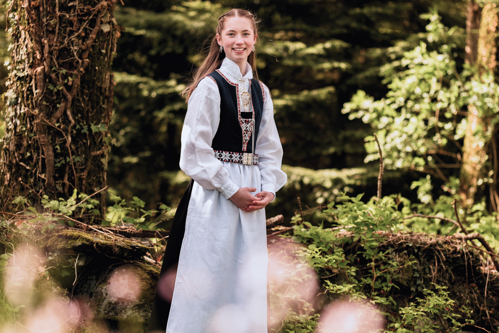 En smilende jente iført tradisjonell norsk folkedrakt står i en skog med grønne trær og planter.