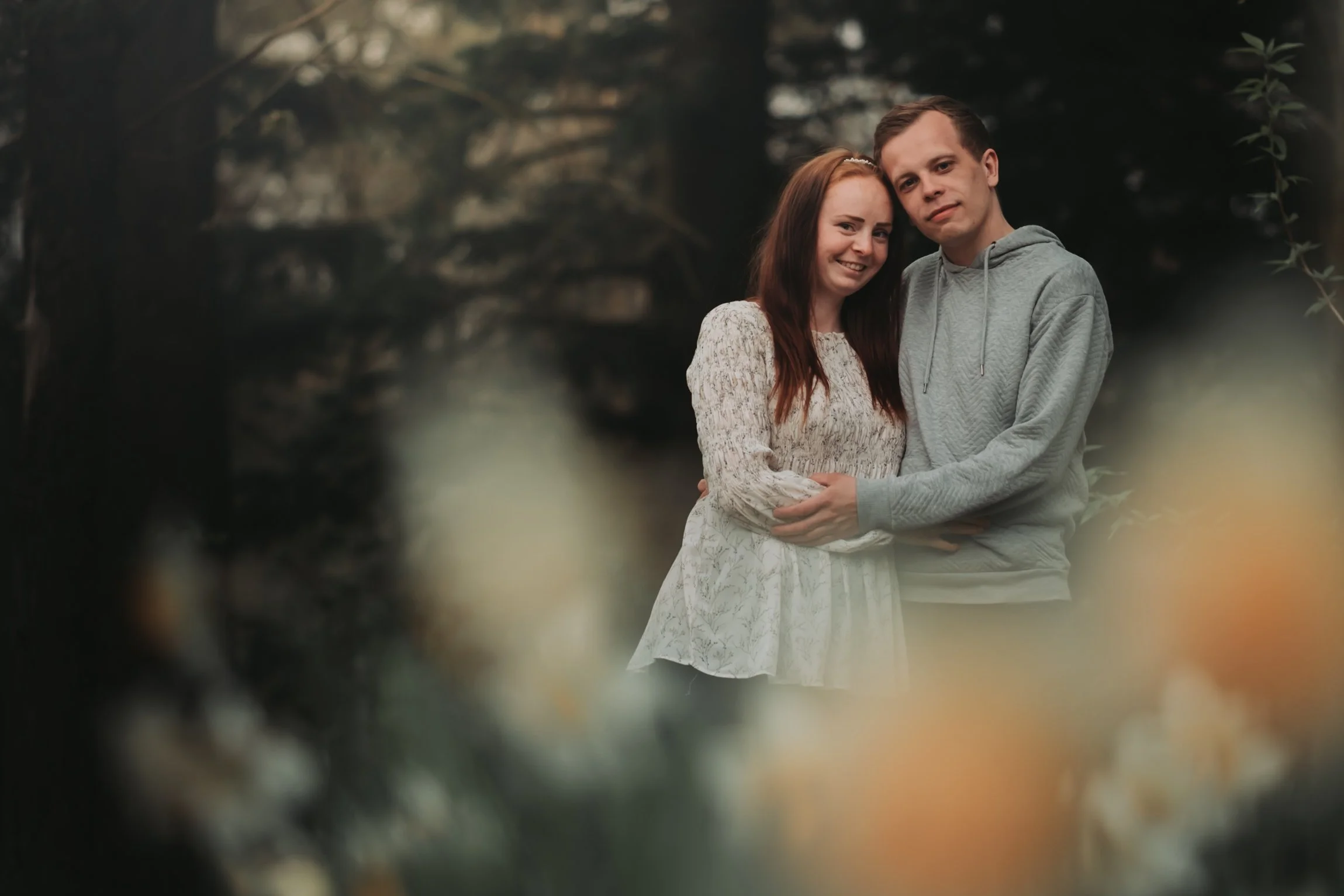 Et ungt par som står sammen utendørs, omgitt av naturlig skjult vegetasjon, med nakne trær i bakgrunnen, smiler og klemmer hverandre.