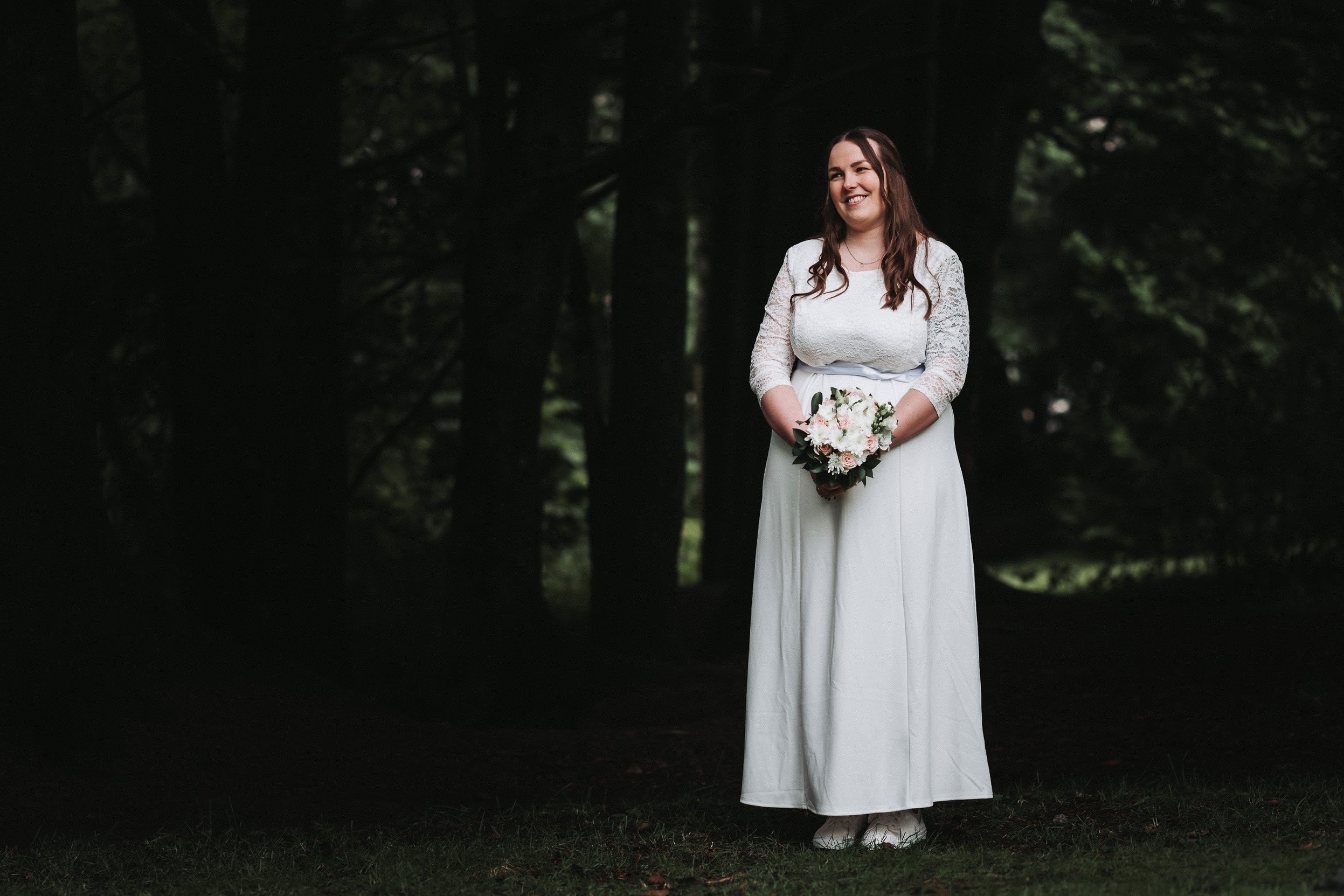 En kvinne i brudekjole holder en blomsterbukett og står i en skog.